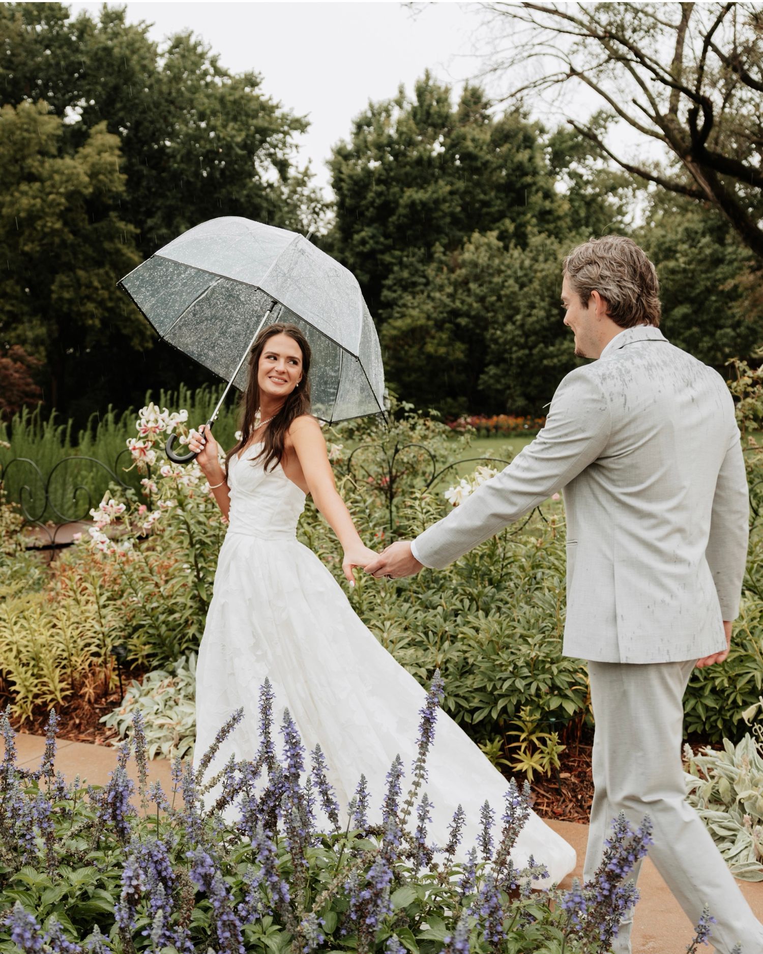 Playful couple with umbrellas dancing in a garden with purple flowers.