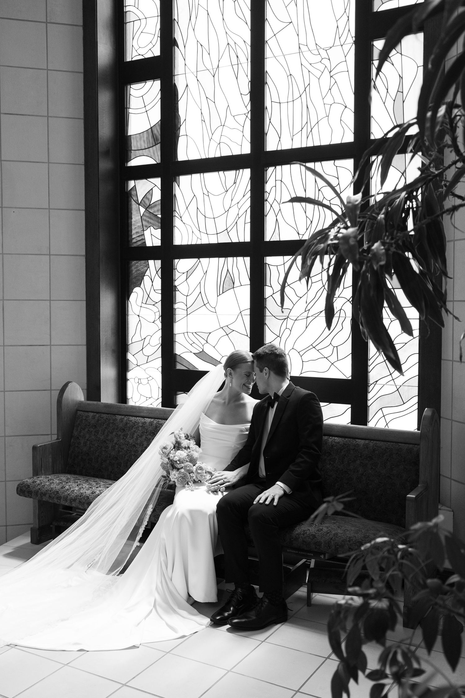 Wedding couple sharing a romantic moment on a couch near large windows.