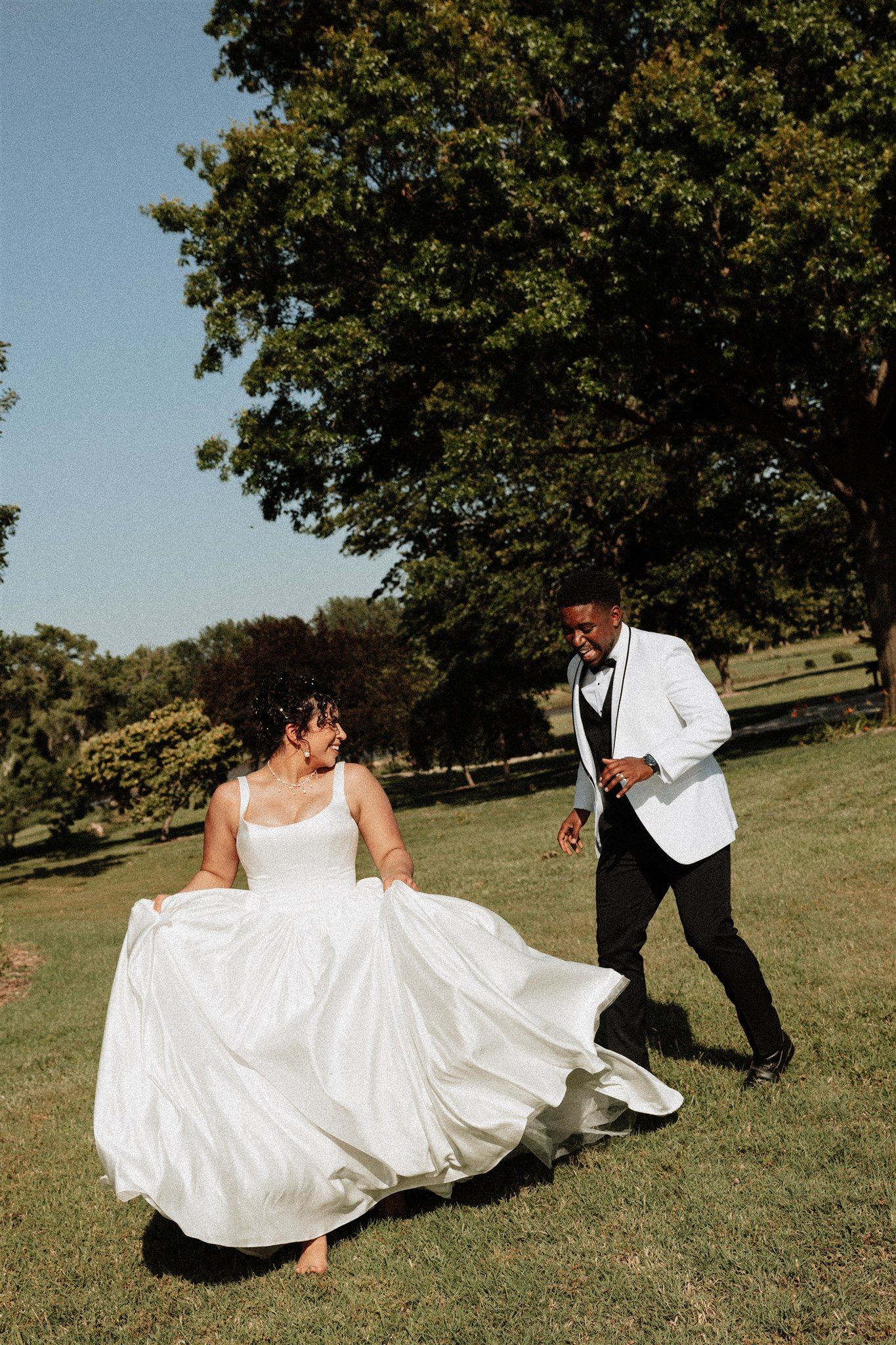 Playful wedding couple running and dancing on grassy hill under trees.