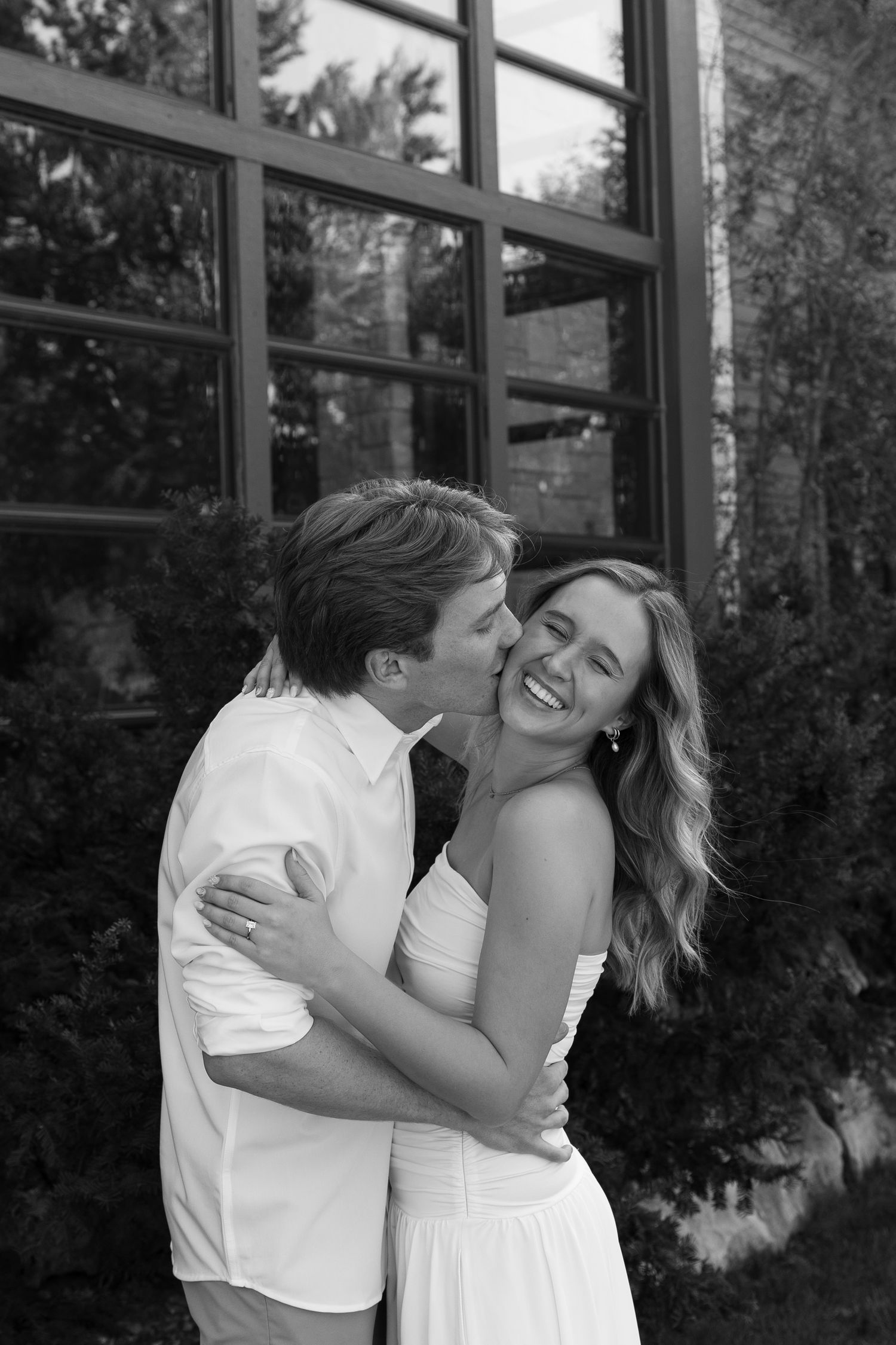 A joyful couple shares a tender moment while laughing together against a window backdrop.