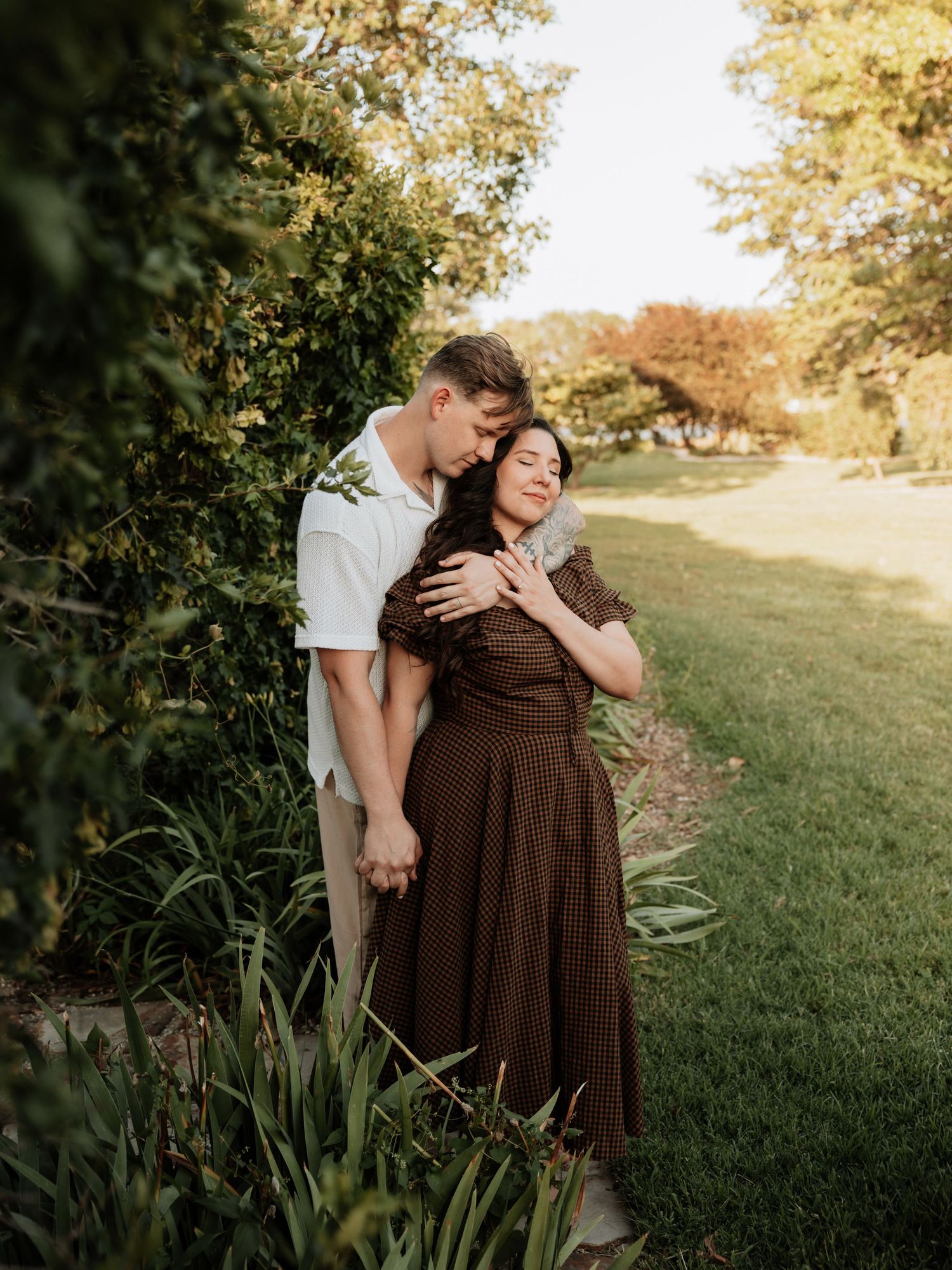 A romantic couple embraces in a garden setting during golden hour with lush greenery in the background.