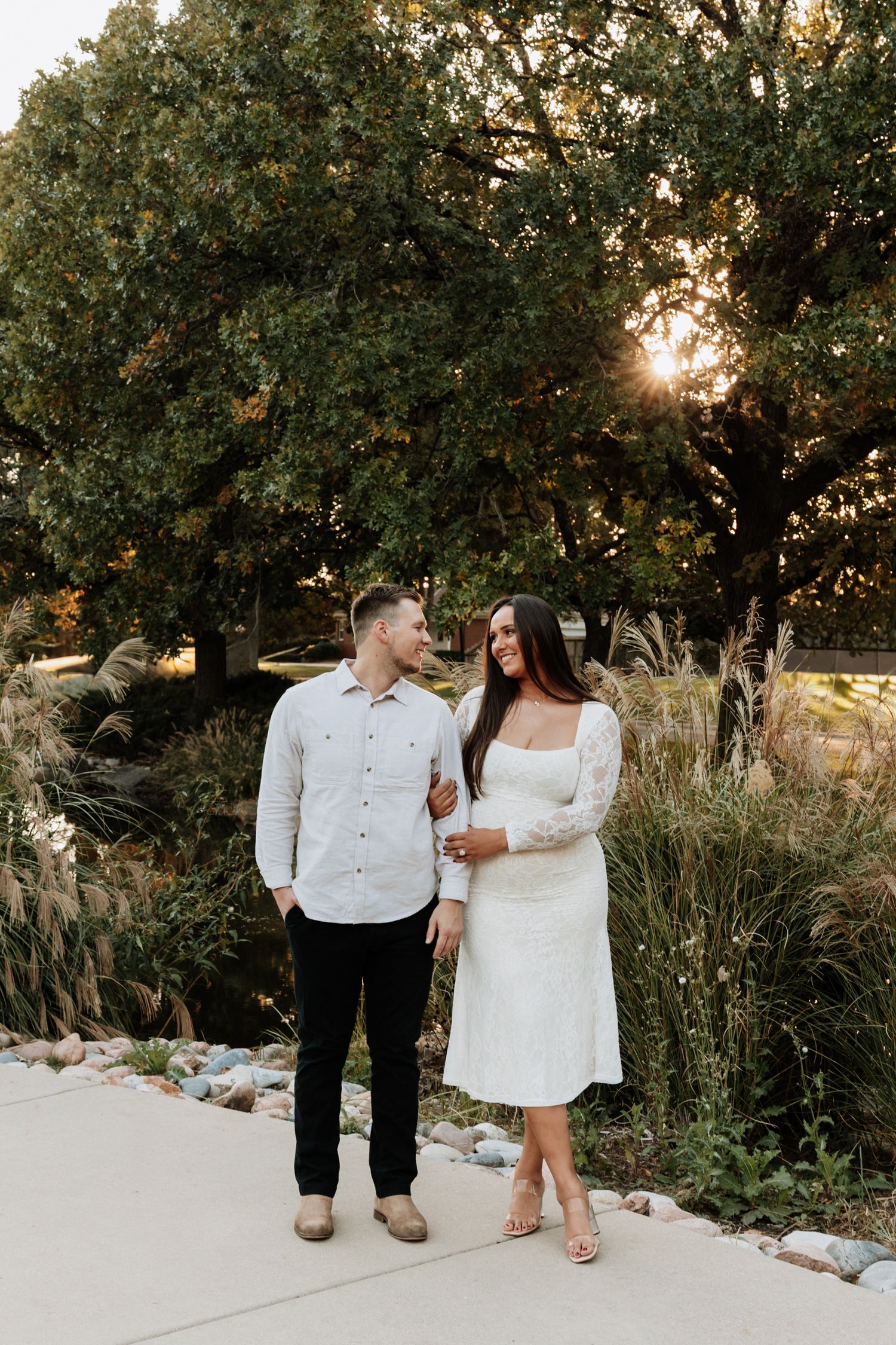 A couple in coordinated white outfits poses together on a garden pathway at sunset.