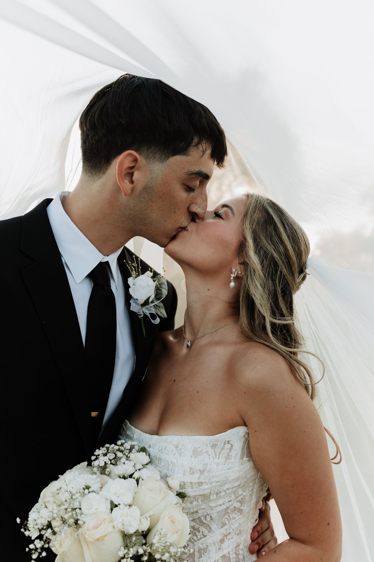 Wedding couple sharing intimate kiss under veil with white bouquet.