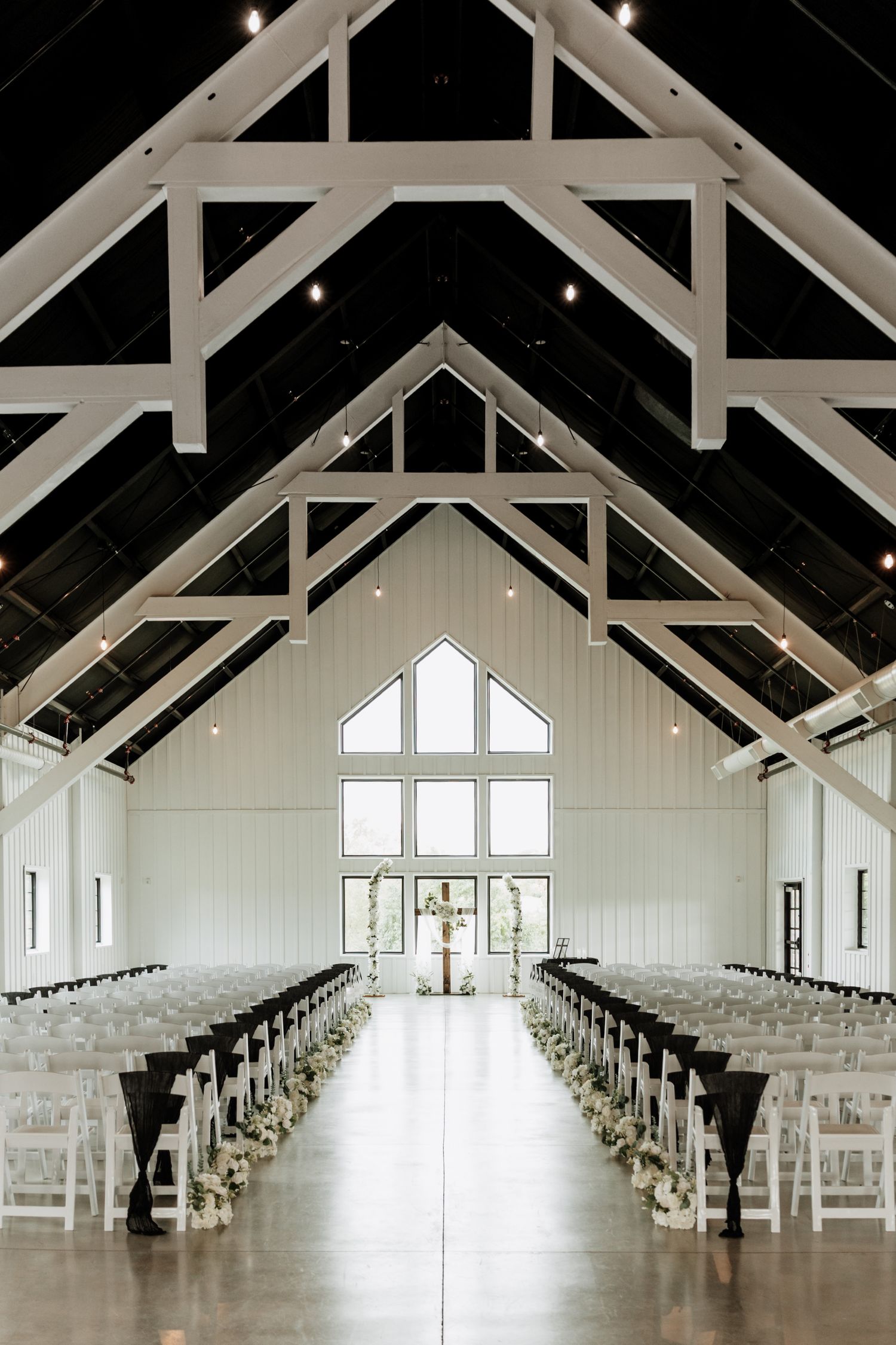 Modern white wedding chapel interior with exposed beams and rows of white chairs leading to large windows.
