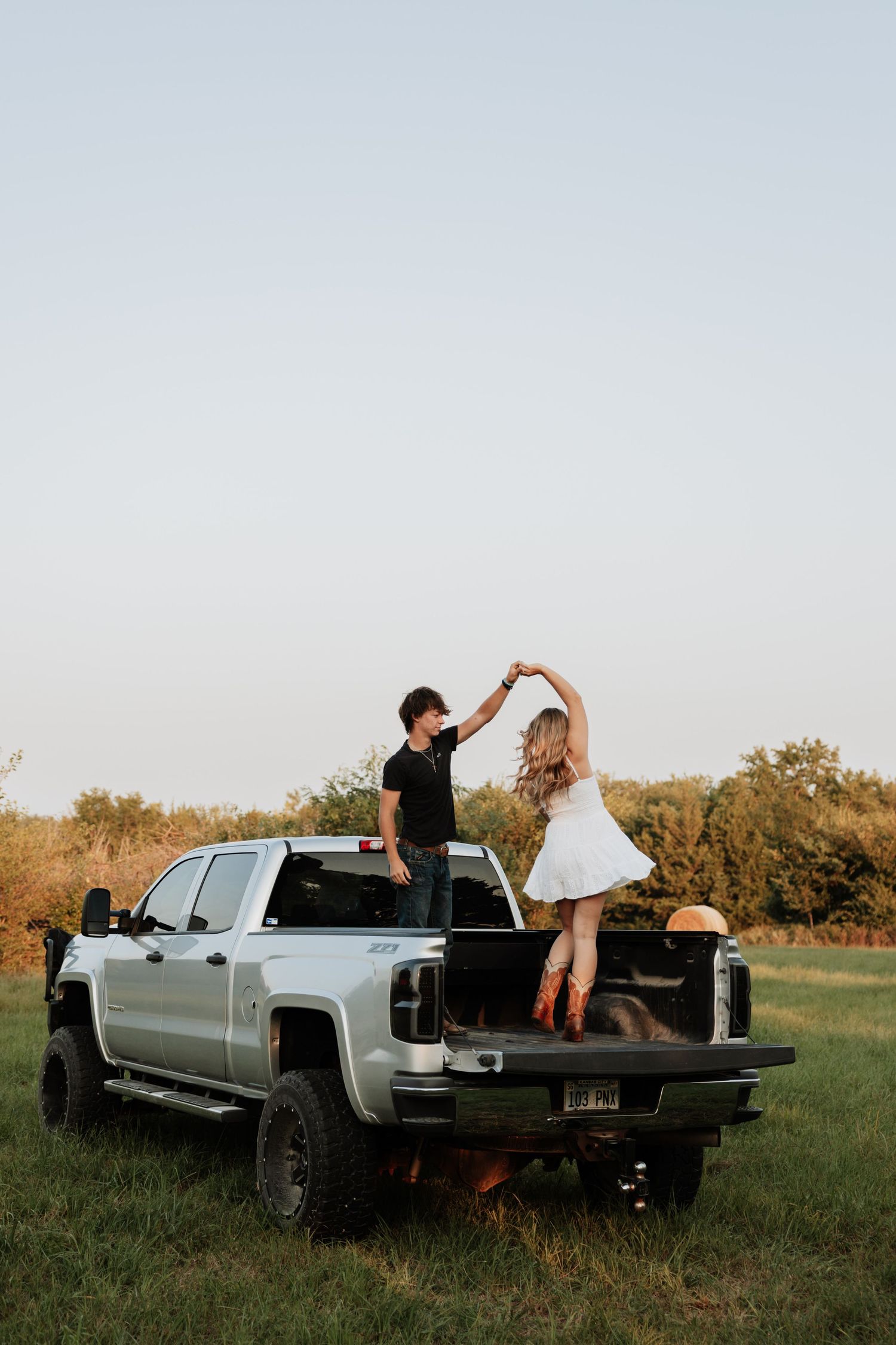 Playful couple dancing on the bed of a silver pickup truck in a field at sunset.