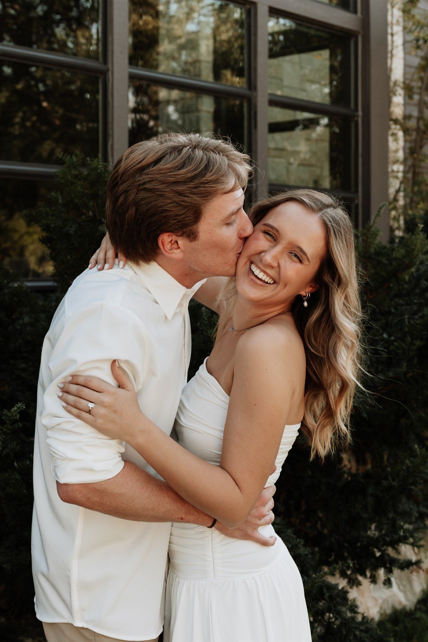 A joyful couple in white outfits embrace and share a tender moment outdoors near a modern building.
