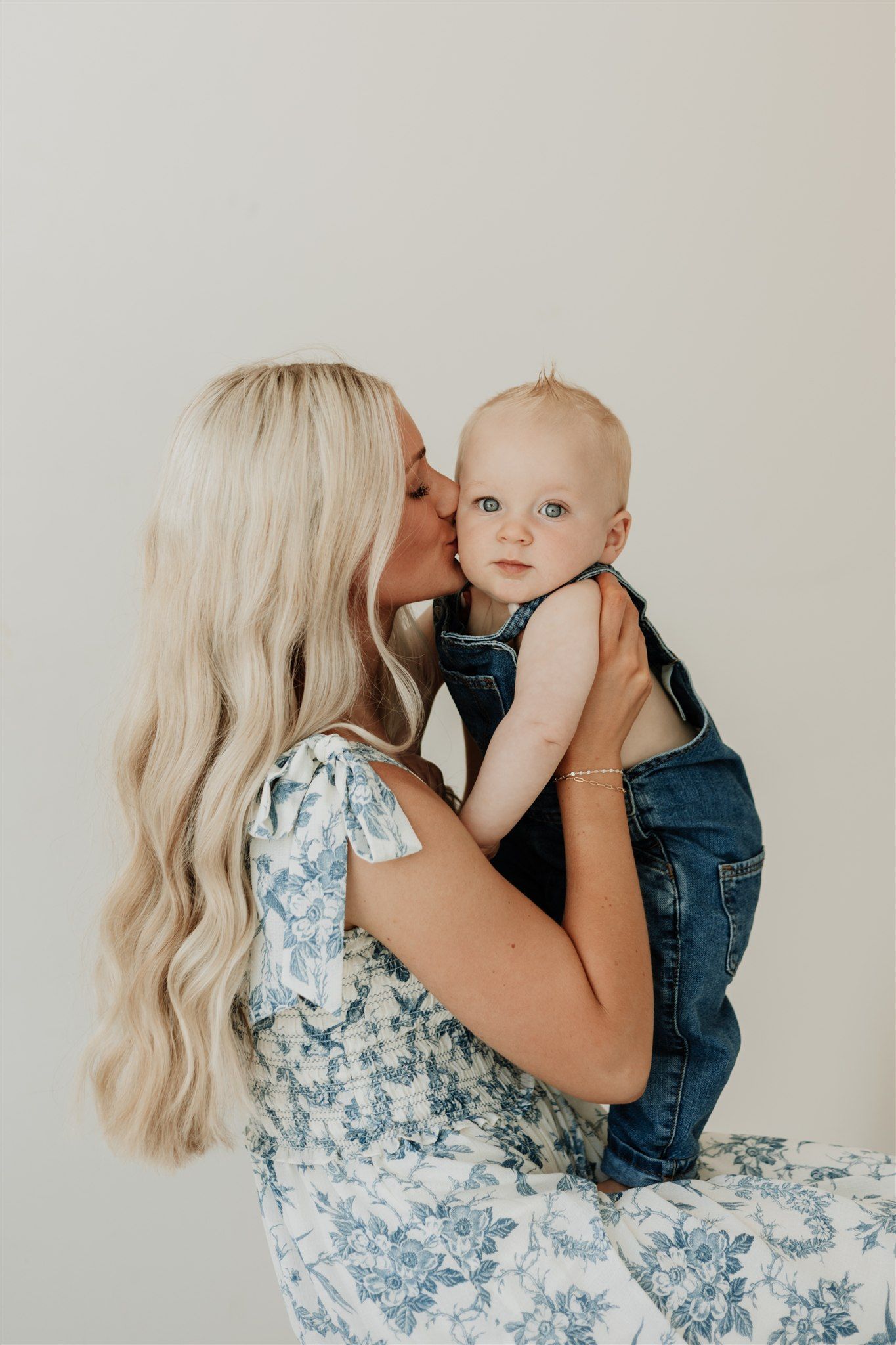 Mother in floral dress holding and kissing baby wearing denim in intimate portrait series.