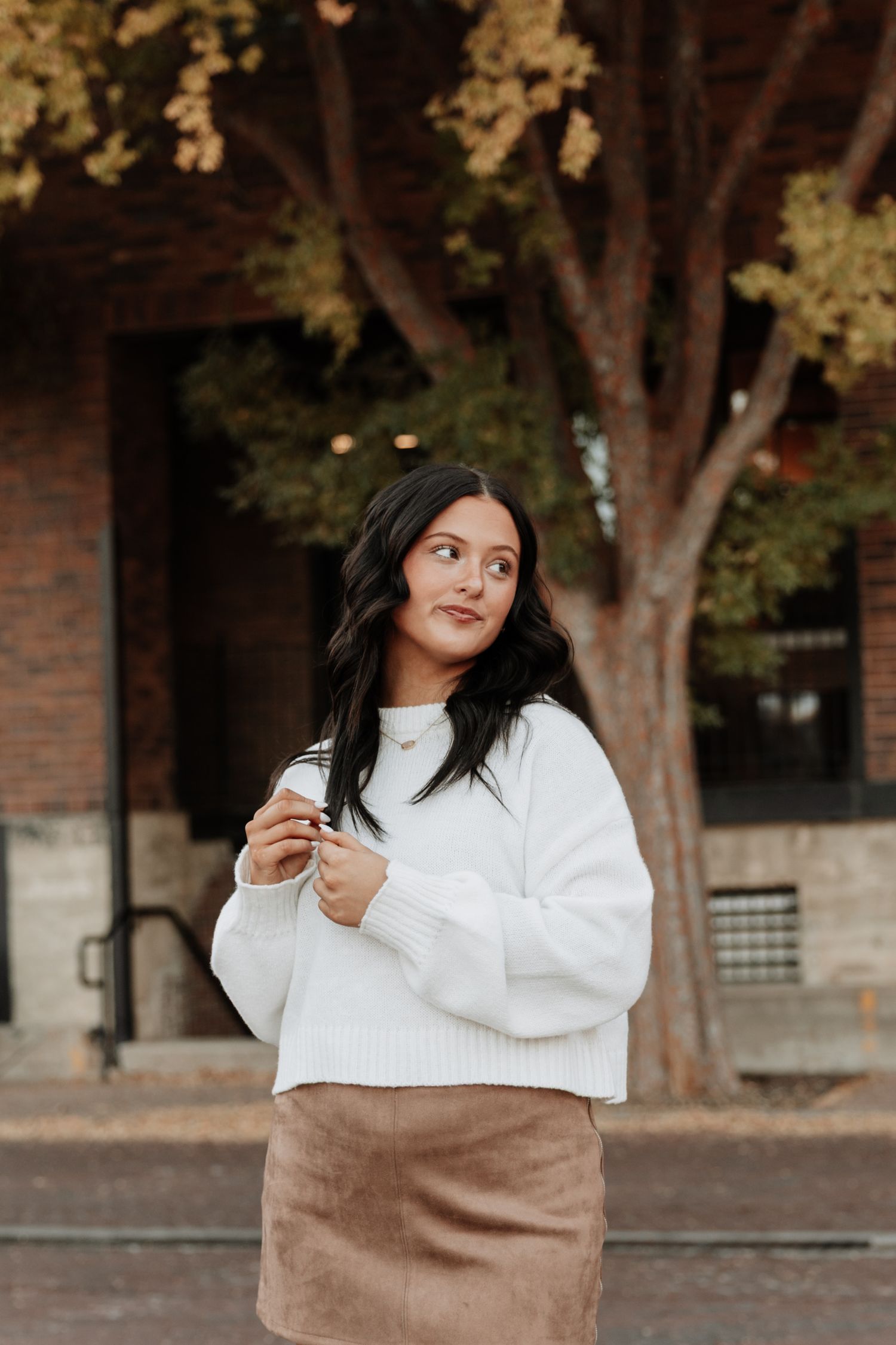 Fashion portrait featuring cozy sweater and tan bag against brick building backdrop.