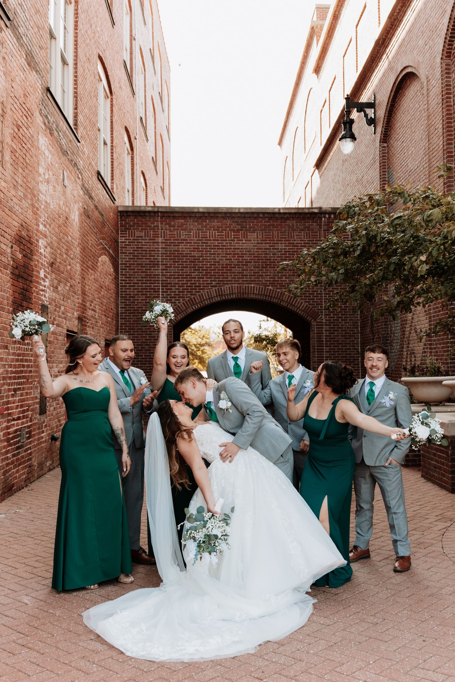 Wedding party in green dresses and gray suits posing against brick wall.