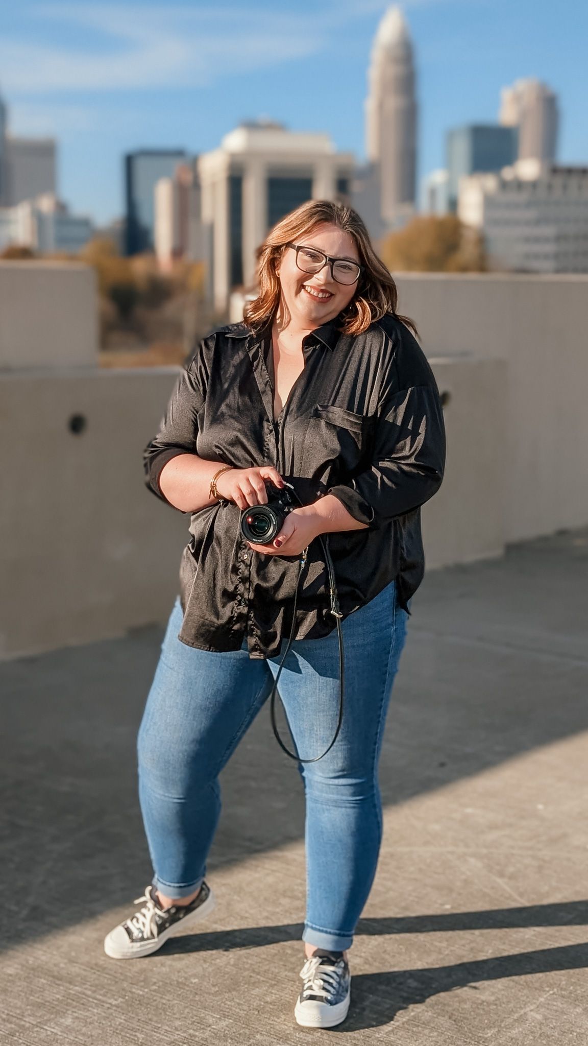 A person in a black silk top and blue jeans poses casually against an urban skyline backdrop.