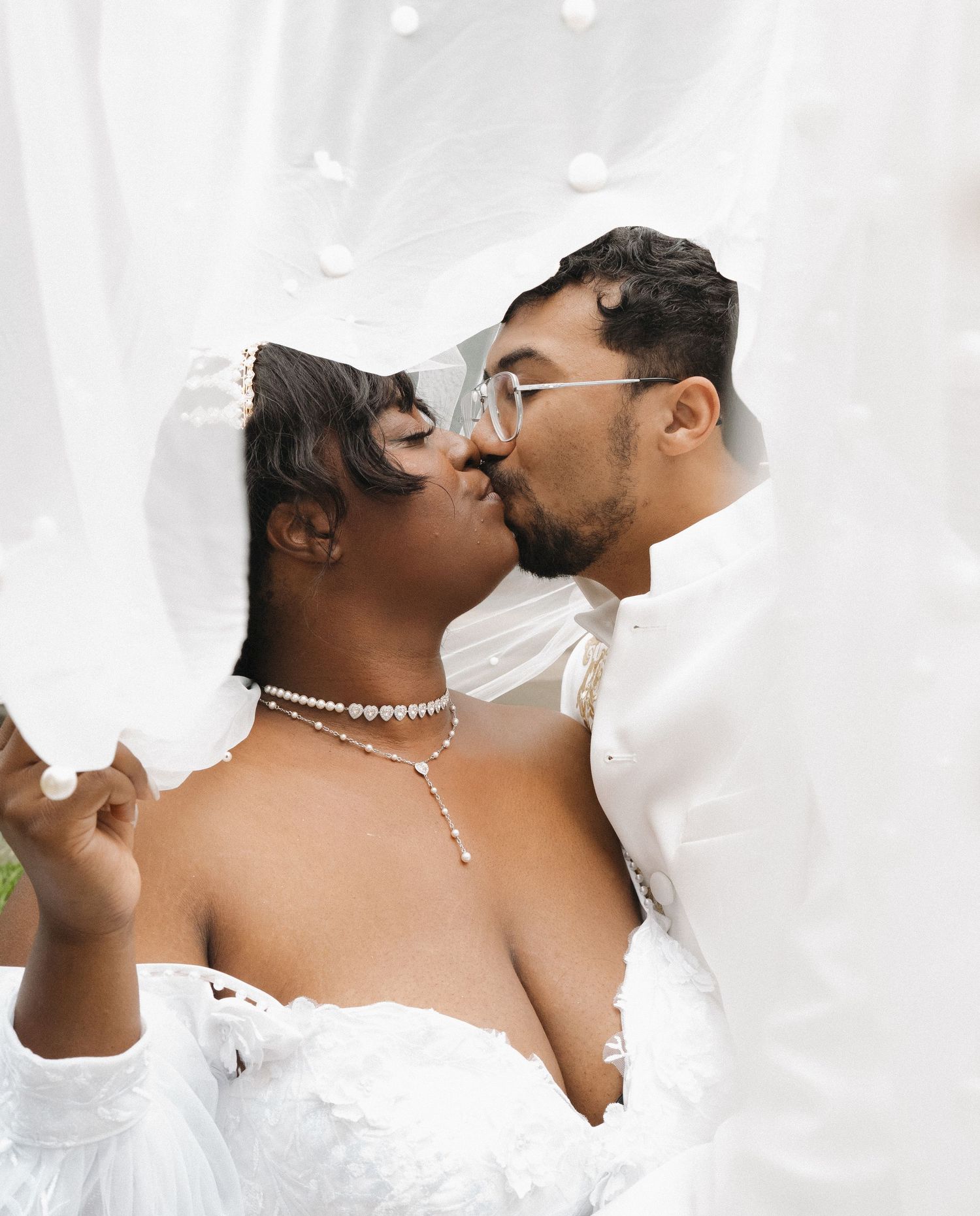 A couple in wedding attire share an intimate kiss while surrounded by soft white veil fabric.