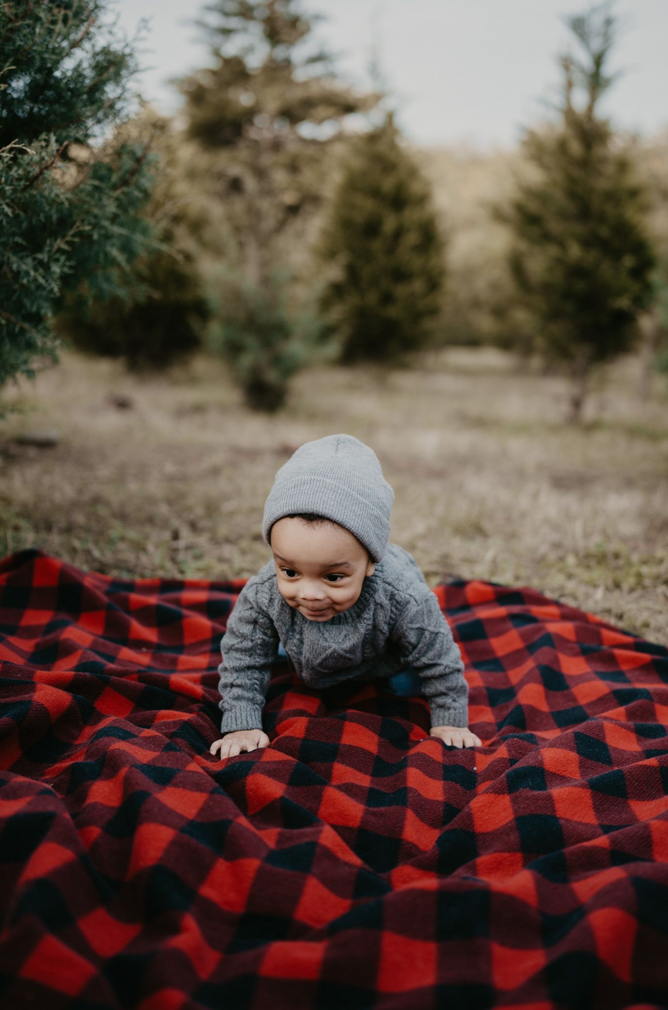 A baby crawls on a red and black buffalo plaid blanket outdoors with evergreen trees in the background.