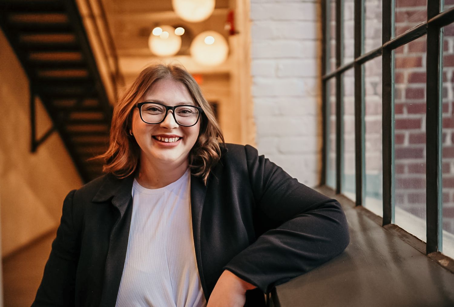 Professional wearing glasses and black blazer sits at desk in warm-lit office setting with brick wall backdrop.
