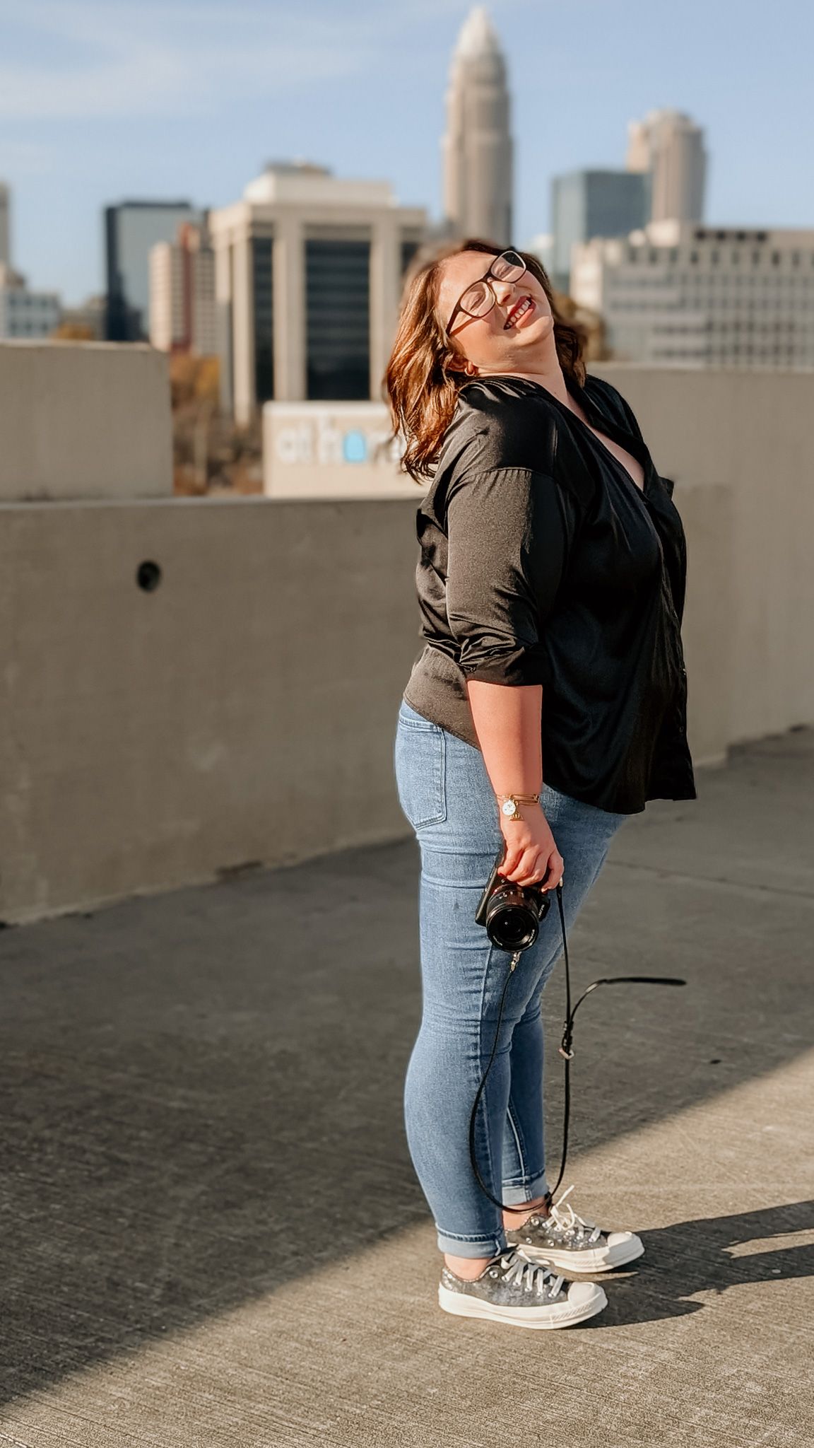 Person in blue jeans and sneakers poses on urban rooftop with city skyline in background.