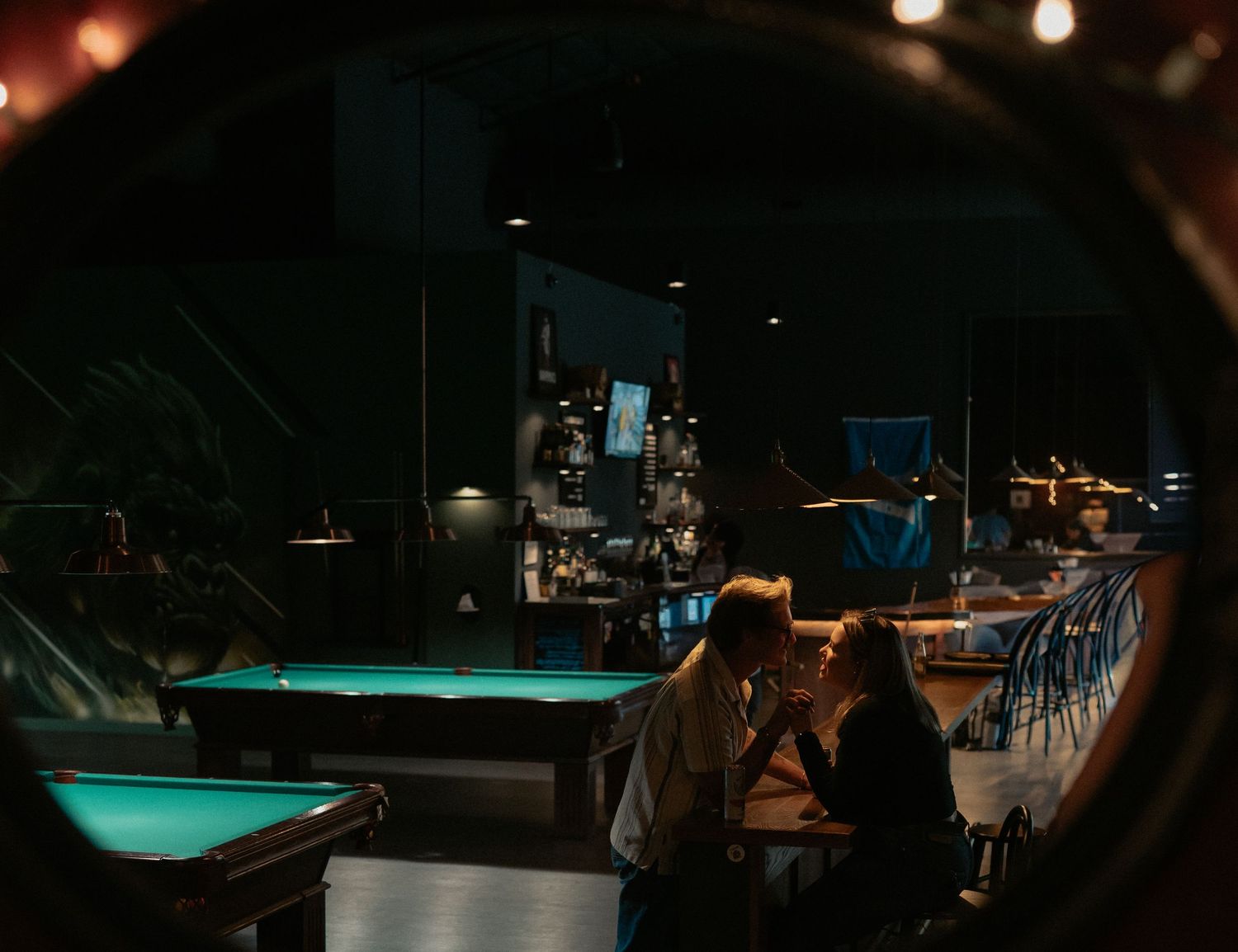A dimly lit bar interior with turquoise pool table lighting and scattered patrons in a moody atmosphere.