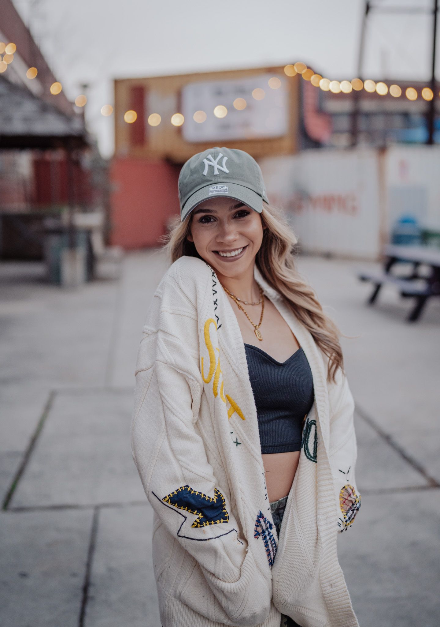 A person wearing a white embroidered jacket and baseball cap poses outdoors with a blurred bokeh background.