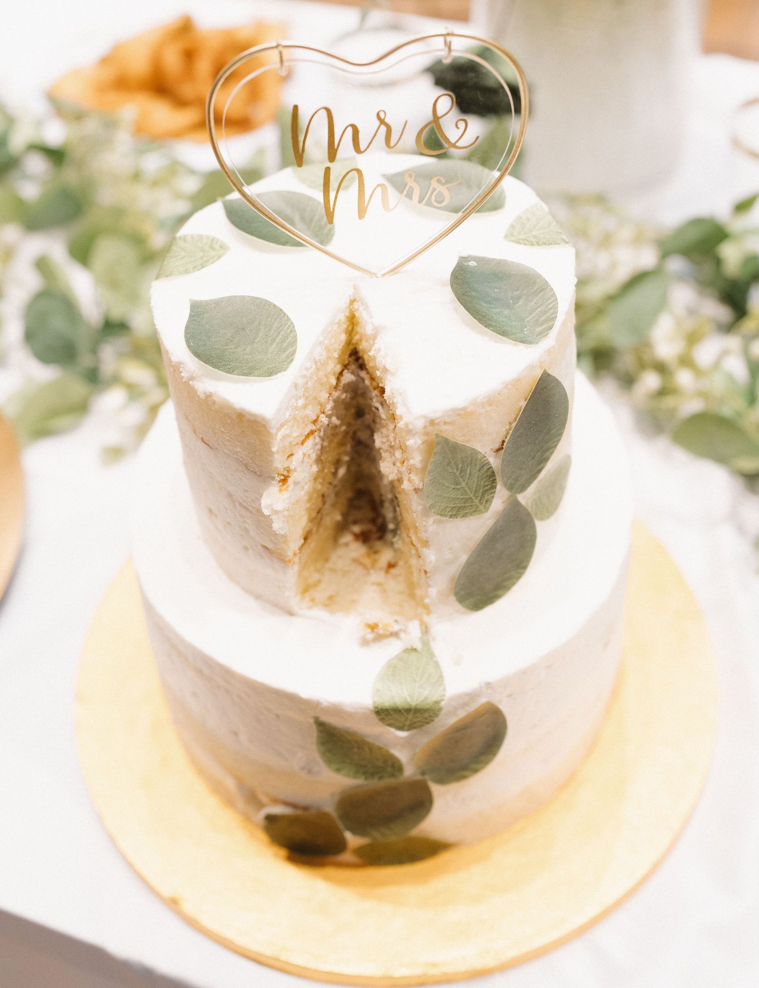 A white wedding cake with eucalyptus leaves displayed in a glass cloche dome on a gold plate.