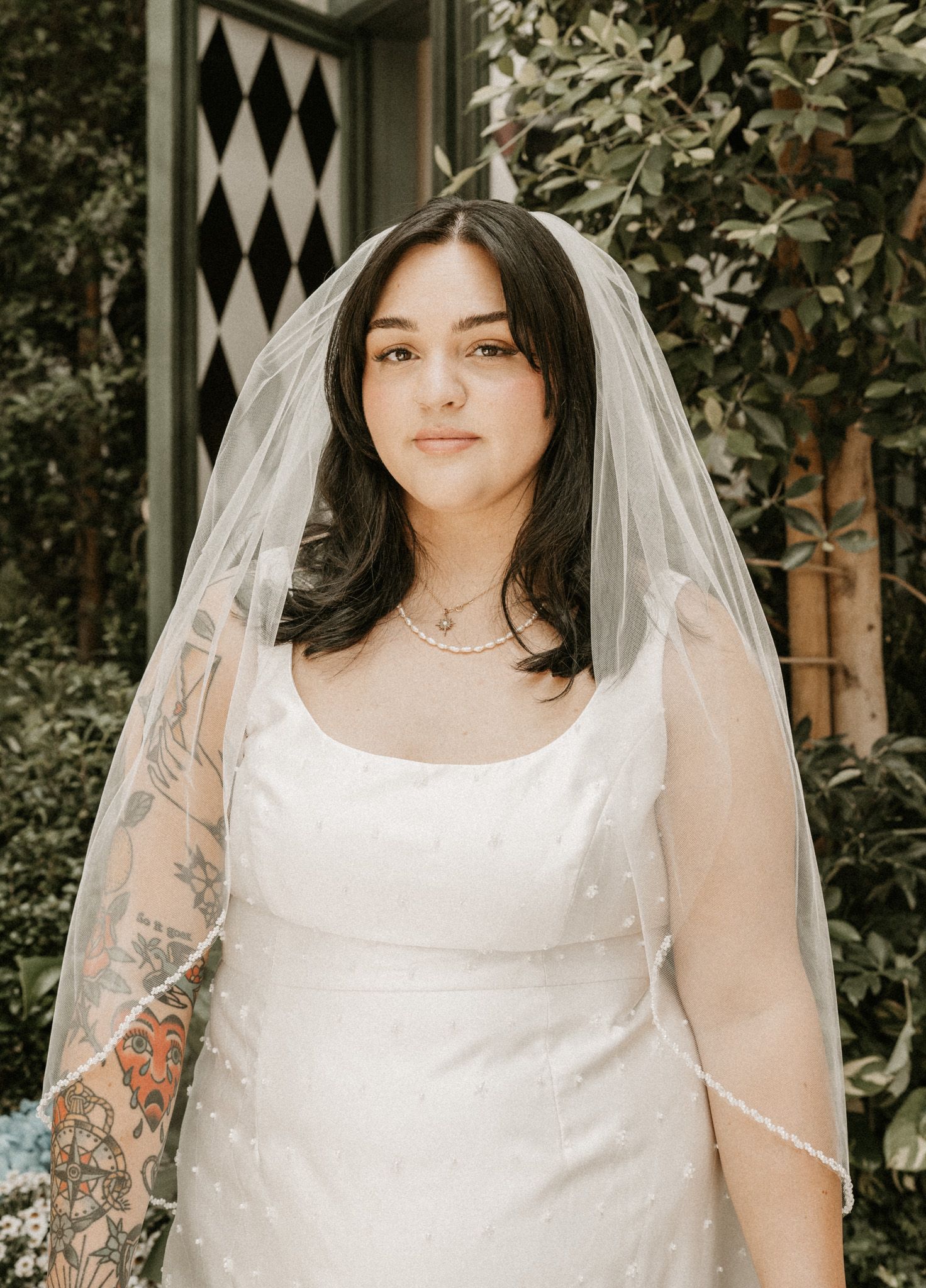 A bride in a sleeveless white wedding dress with tattoos poses in front of a black and white patterned backdrop.