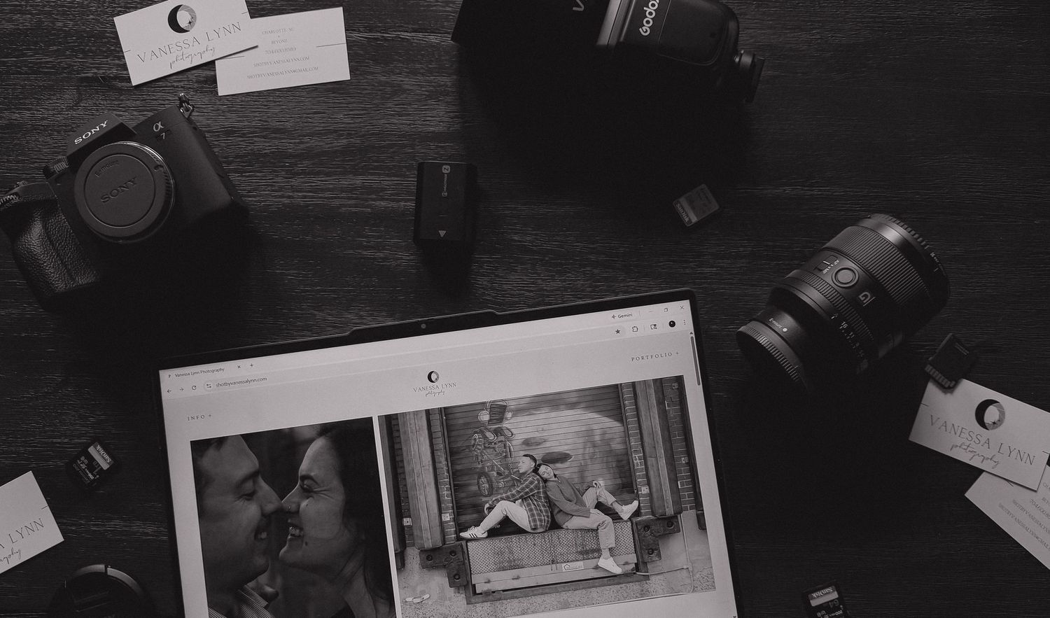 Black and white photograph of vintage photographs and camera equipment arranged on a dark wooden surface.