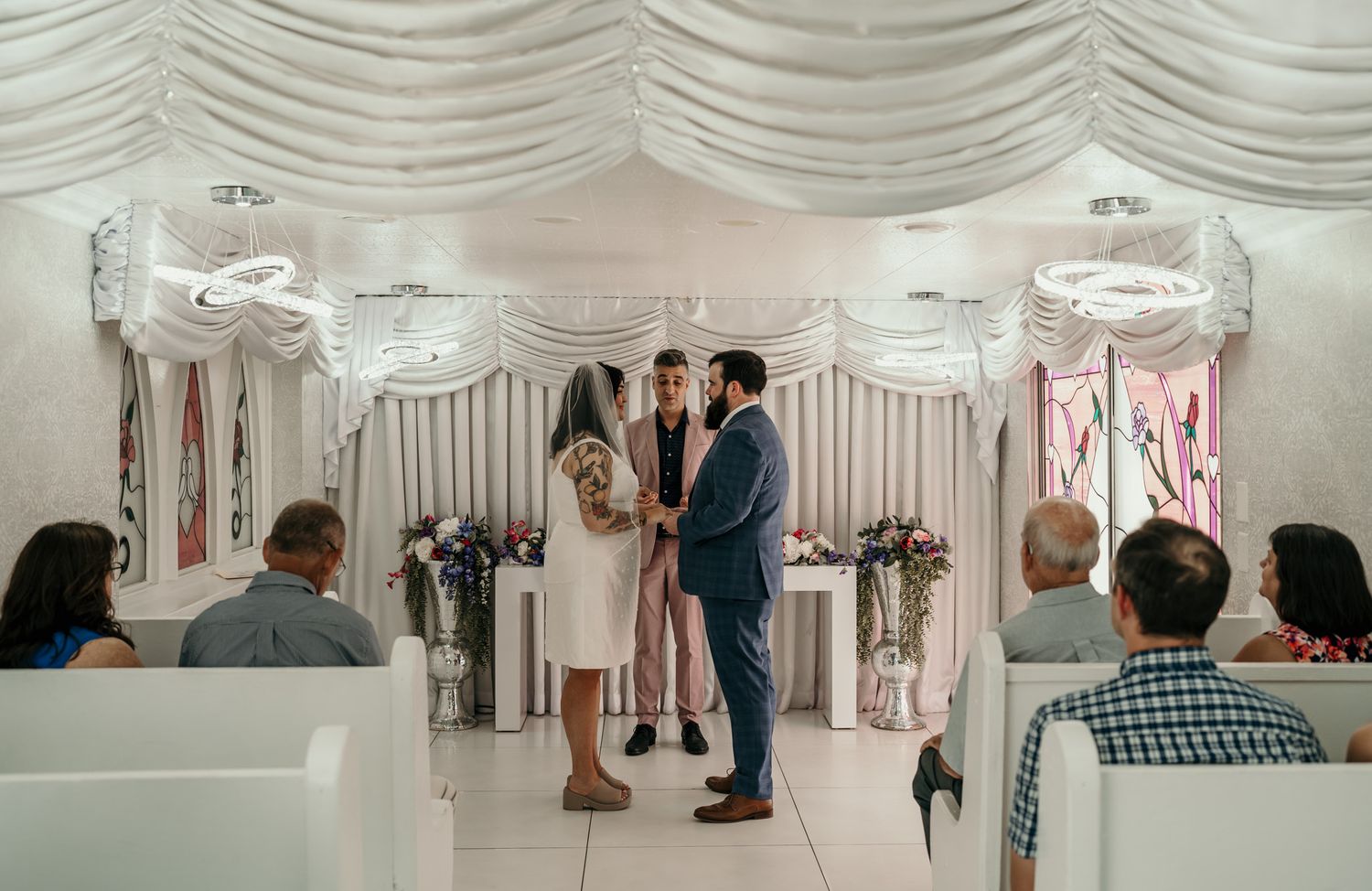 Couple exchanging vows at a small wedding chapel ceremony with white draped decor and seated guests.