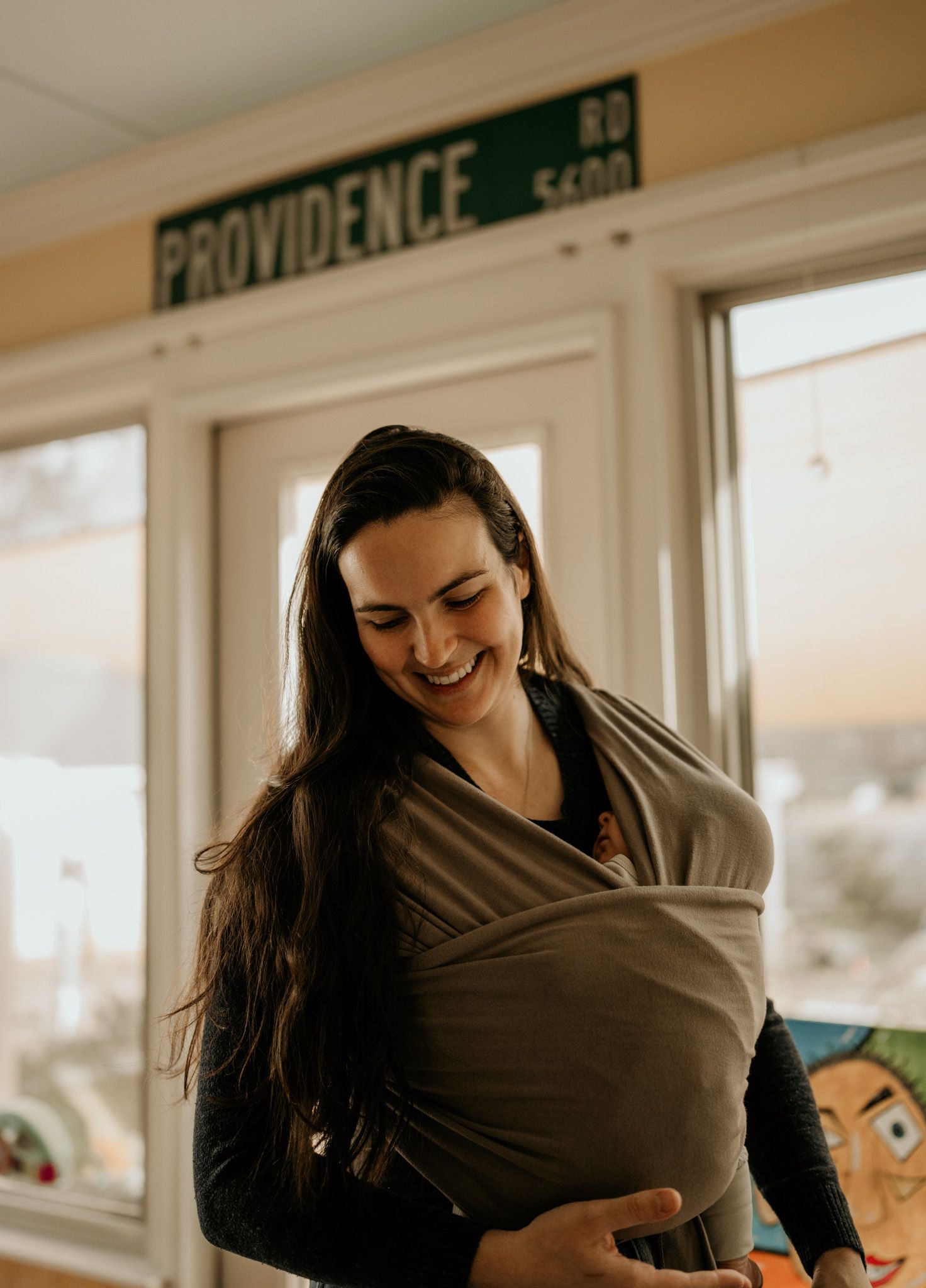 A person wearing a brown baby carrier wrap smiles while holding an infant in different poses against a sunlit window.