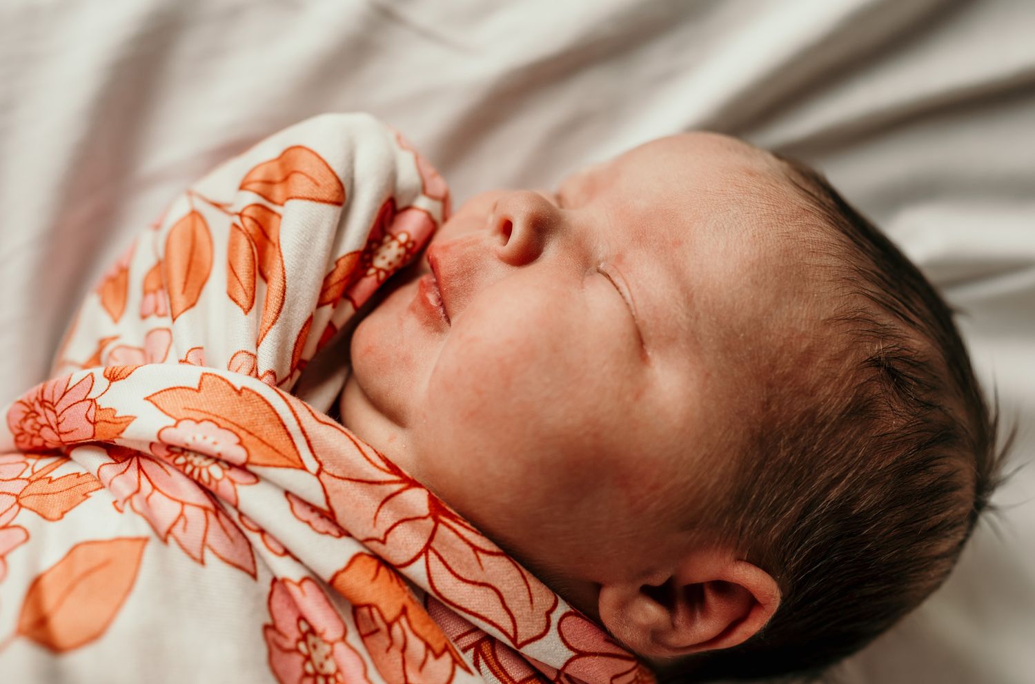 A sleeping baby wearing orange floral pajamas rests peacefully on white bedding.