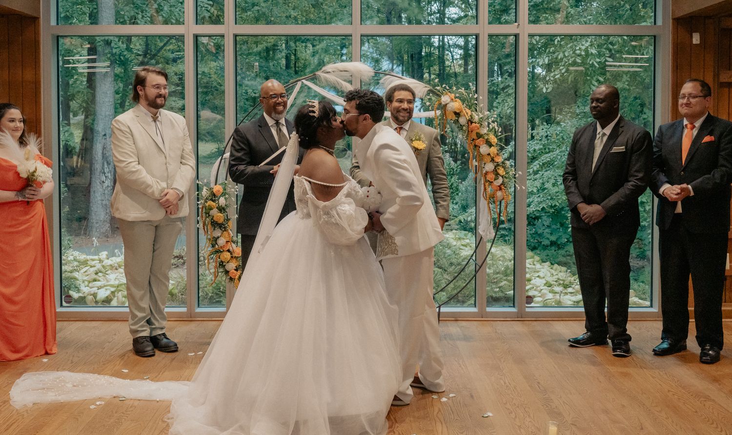 A couple shares their first kiss at a wedding reception in an elegant venue with large windows and watching guests.