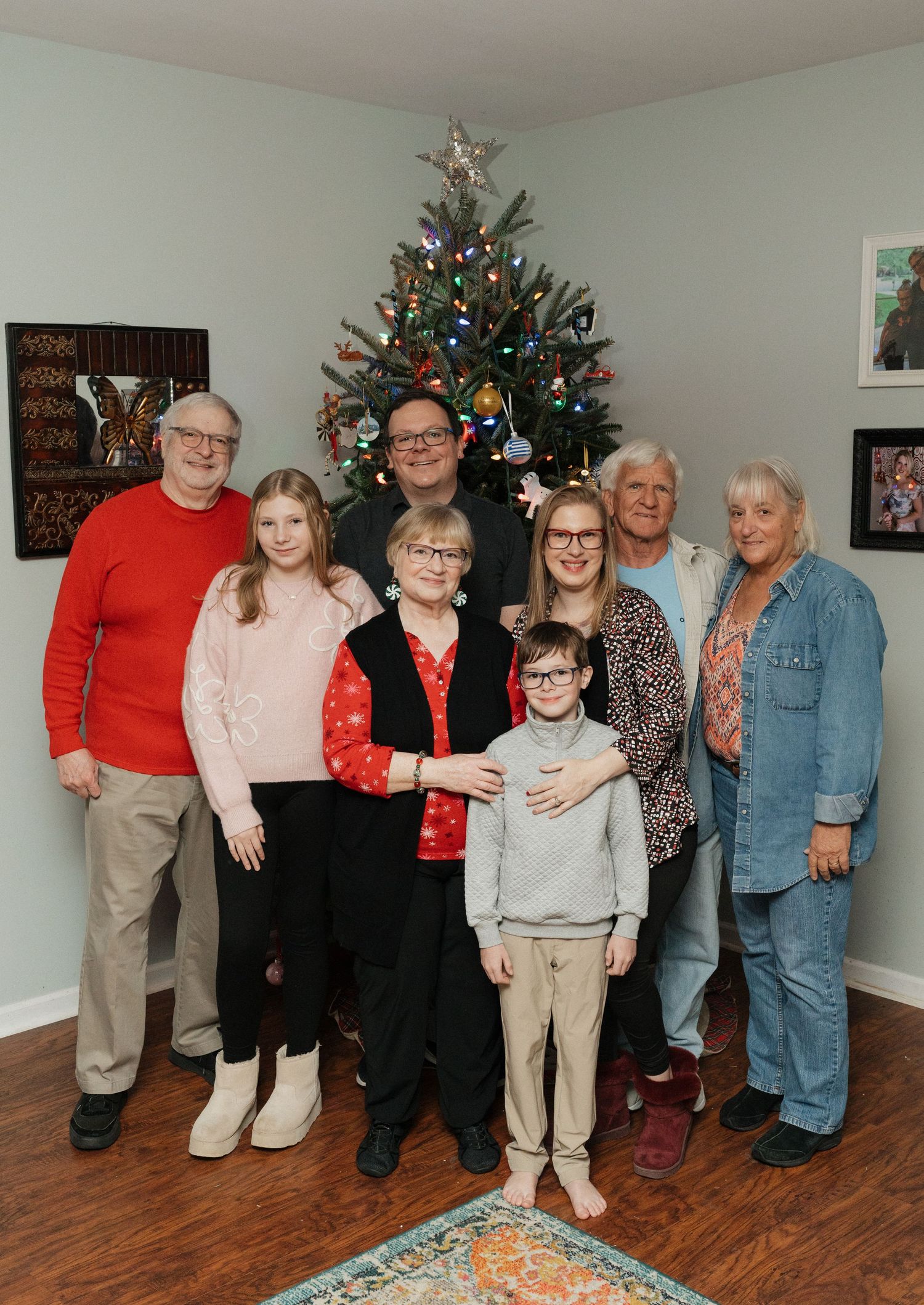 A group of people gather around a decorated Christmas tree in a living room with hardwood floors.