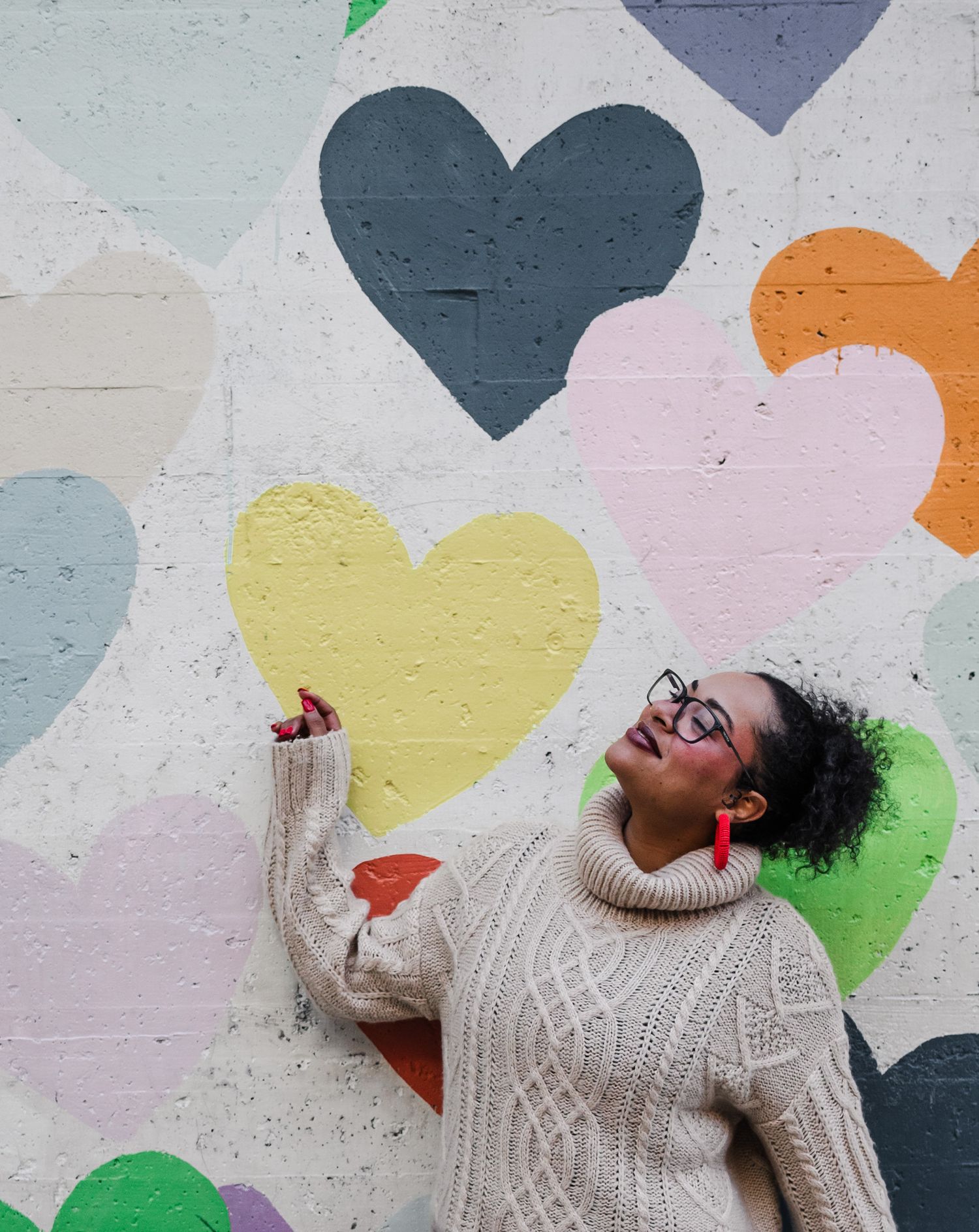 A person in a cream cable knit sweater poses joyfully against a colorful wall decorated with pastel heart shapes.