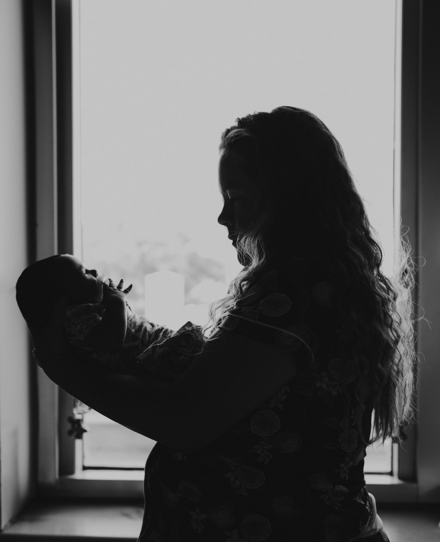 A silhouetted figure stands by a window holding a baby, creating a tender black and white moment in natural light.