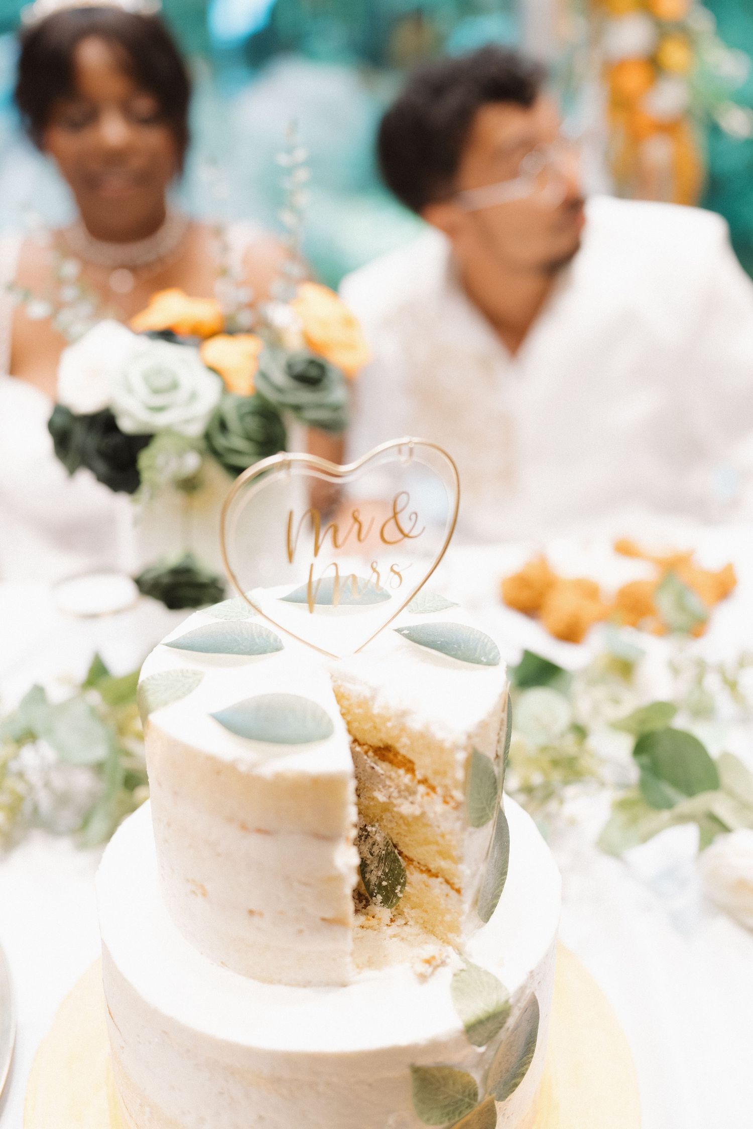 White wedding cake decorated with eucalyptus leaves and gold accents sits on an elegant table at a reception.