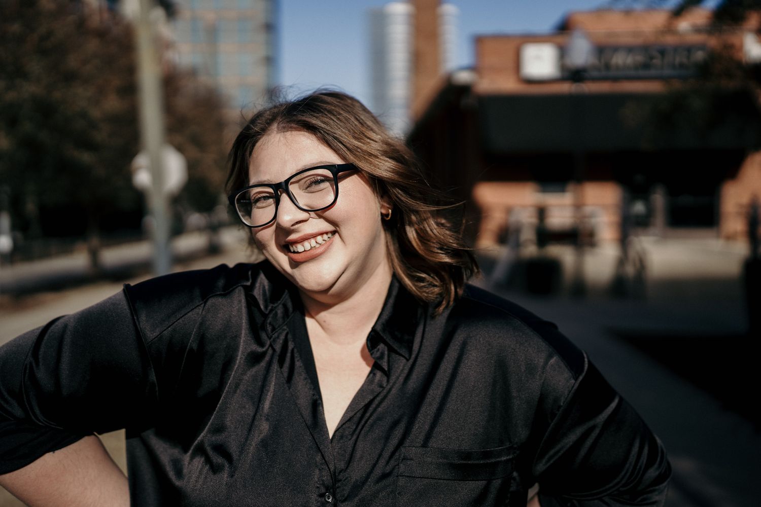 A joyful person wearing glasses and a black button-down shirt smiles warmly on a sunny urban street.