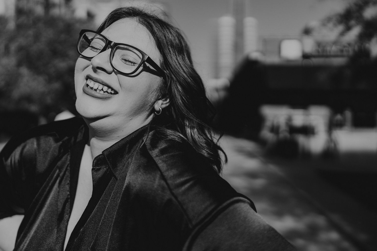 Black and white portrait of someone laughing joyfully while wearing glasses and a leather jacket outdoors.