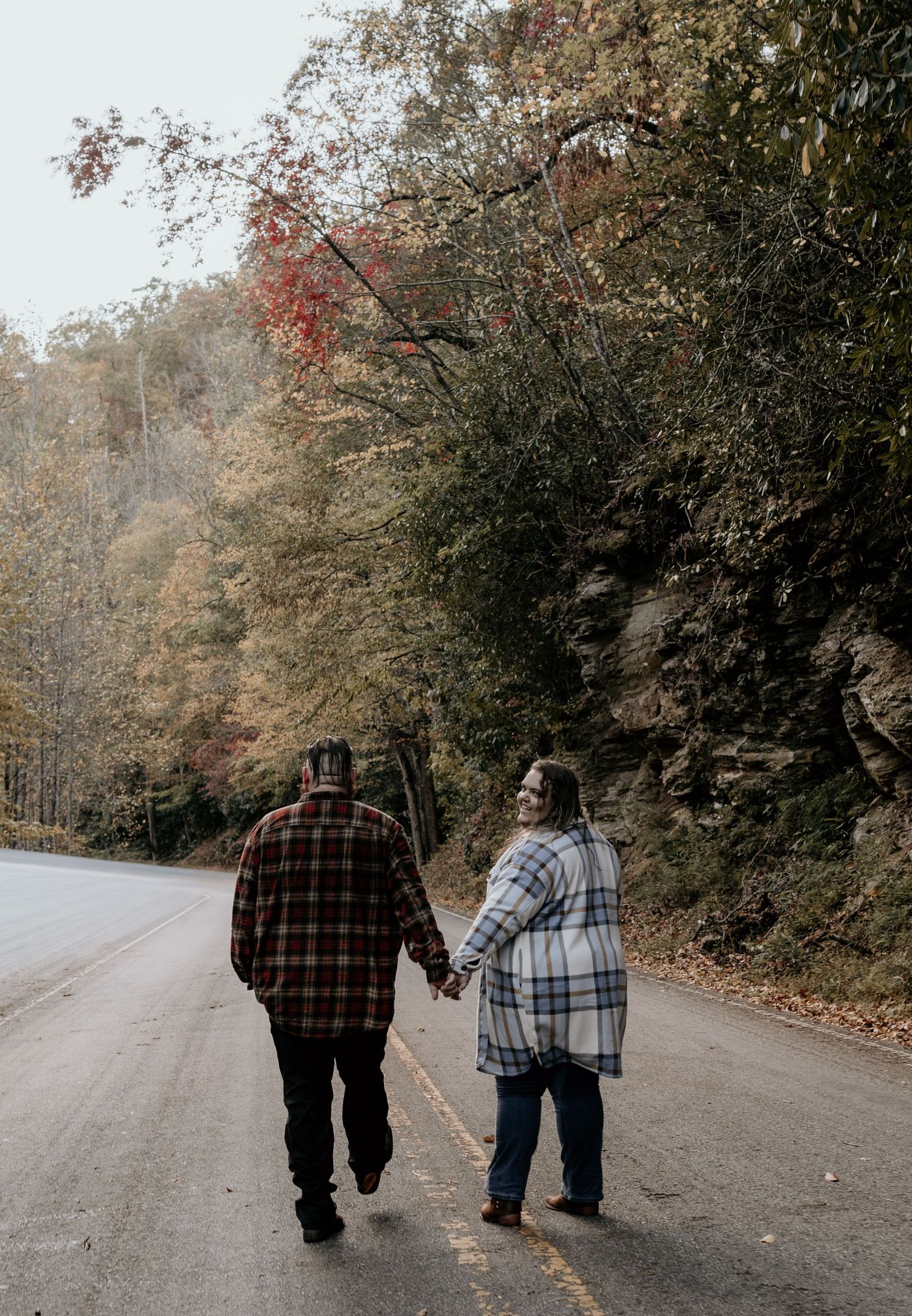 Two people walk together down a winding autumn road surrounded by trees with changing fall foliage.