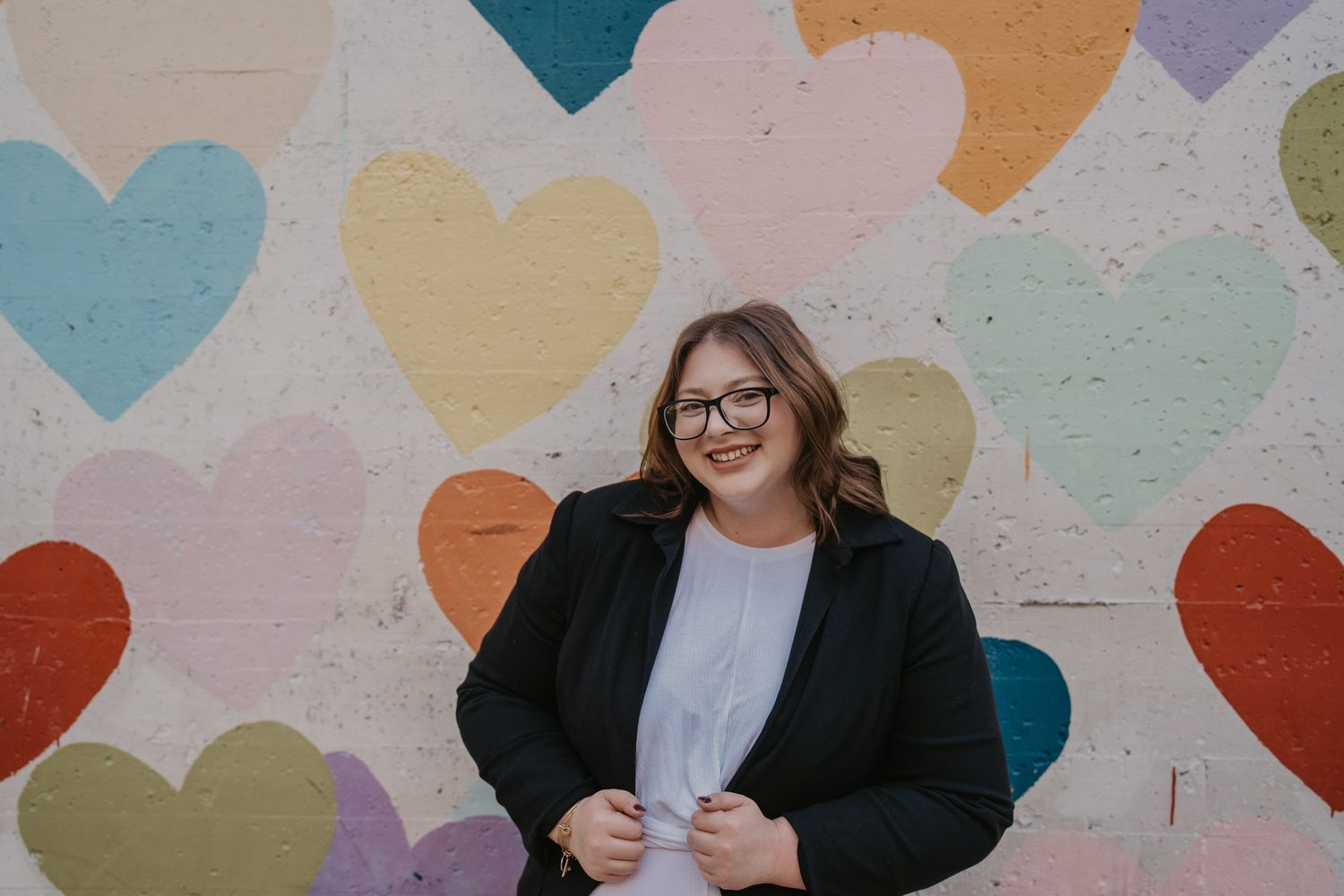 A person in a black blazer stands against a colorful heart-patterned wall for a photo.