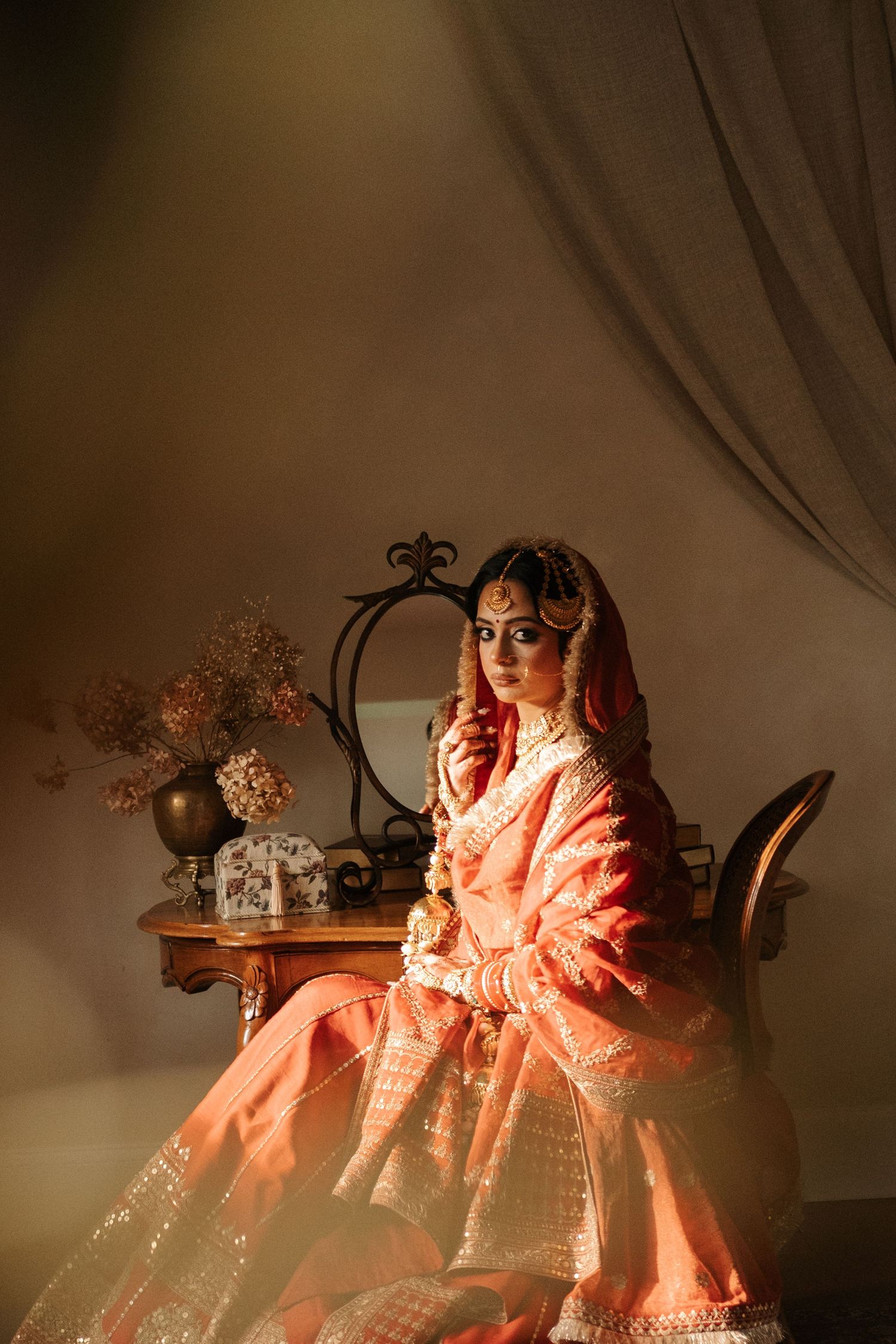 Indian bride in peach lehenga seated at vintage vanity table with dramatic lighting and gold bridal jewelry.