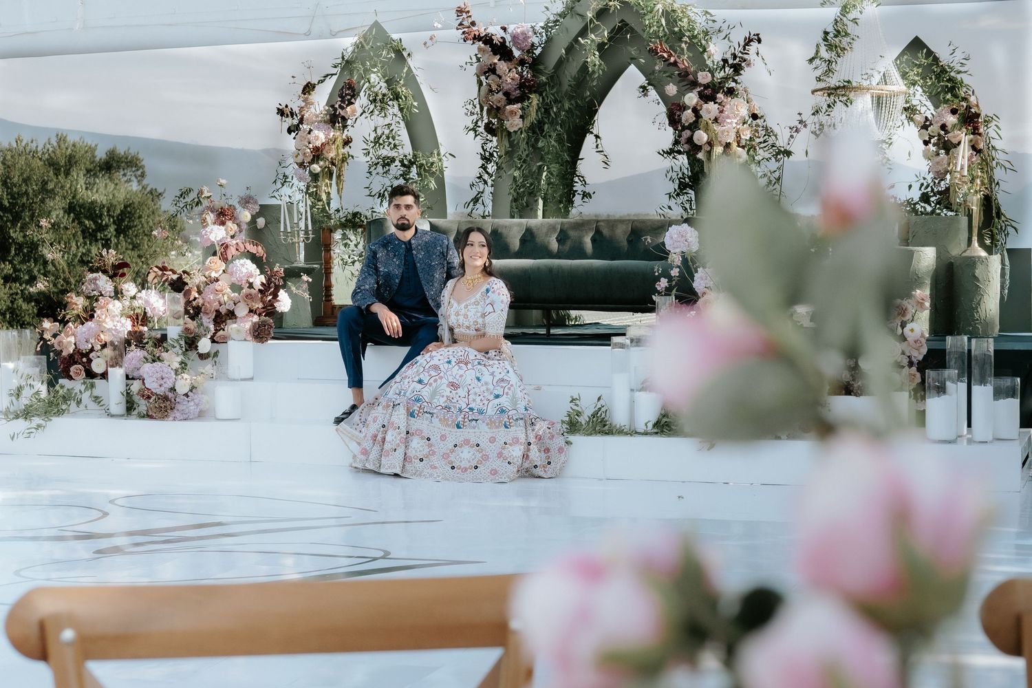 A couple poses on a white marble floor surrounded by gothic floral arches and soft pink roses at a wedding.