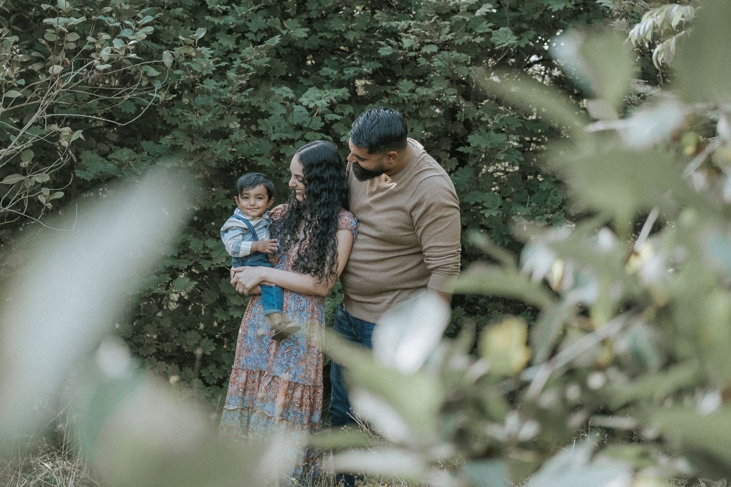 Family of three framed by lush green foliage, mother holding toddler while parents share tender moment.