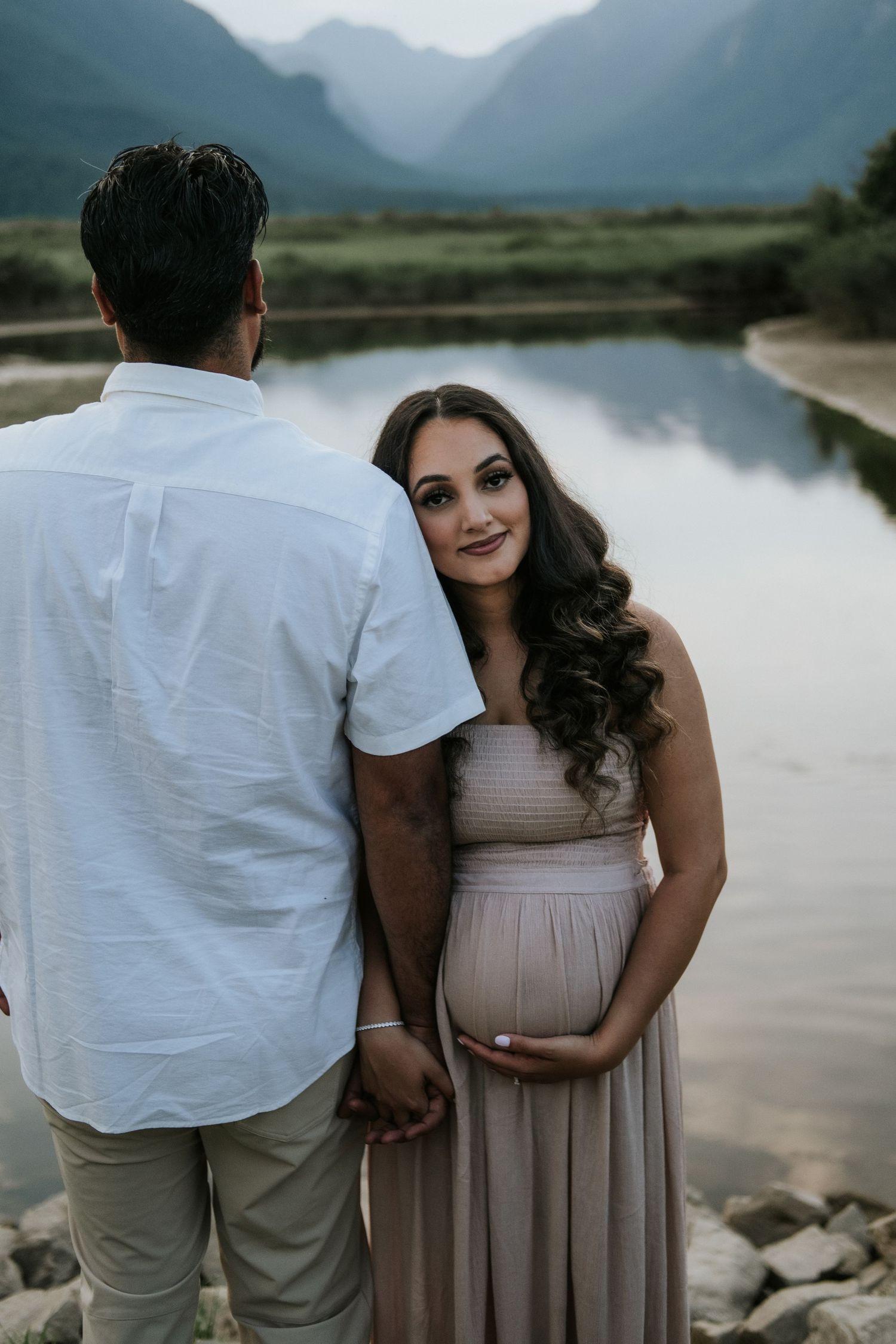 Pregnant woman in blush gown smiling over shoulder while holding partner's hand near a mountain lake.