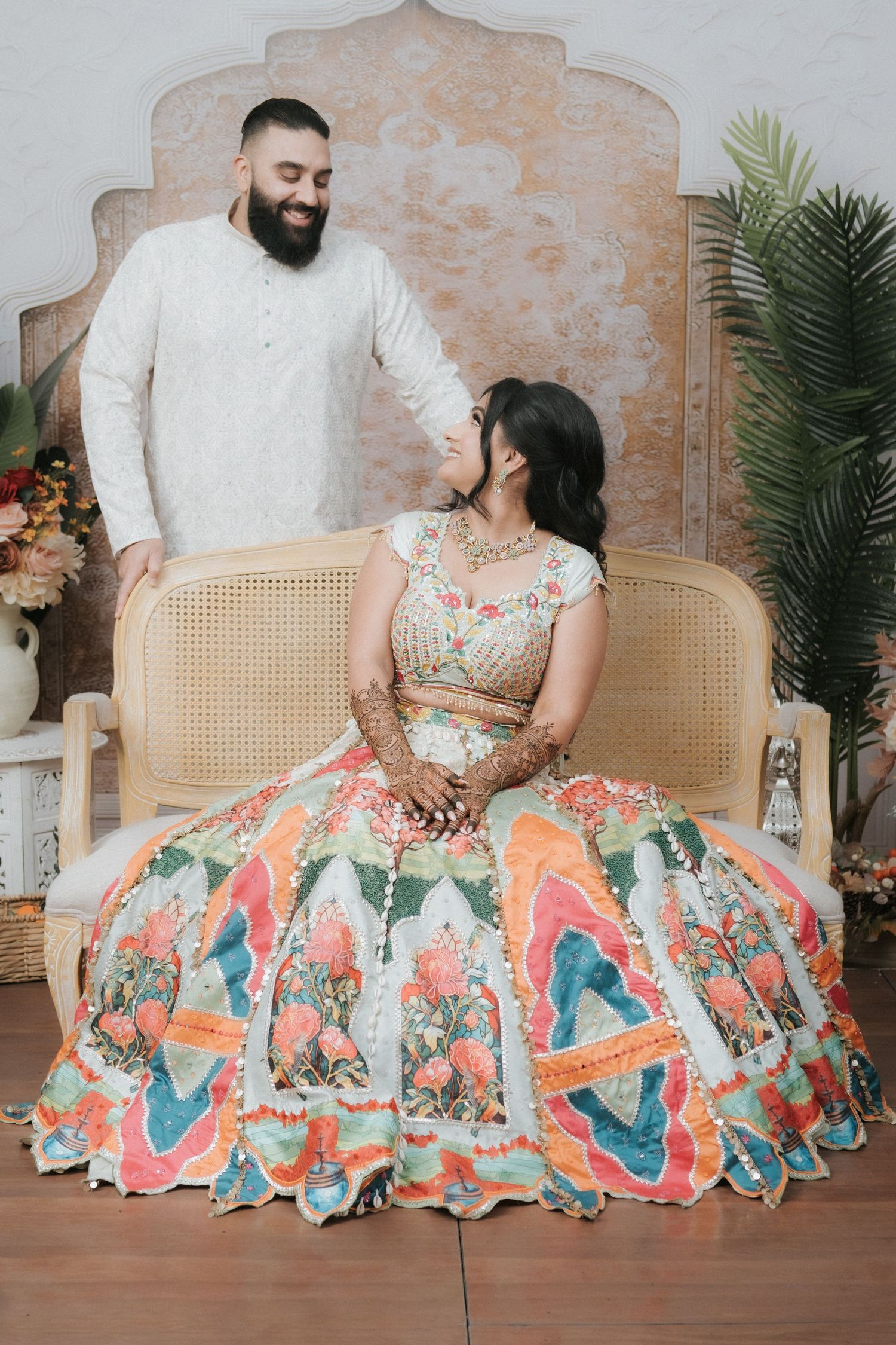 Groom in white kurta smiling at seated bride wearing colorful floral lehenga with intricate mehndi designs.