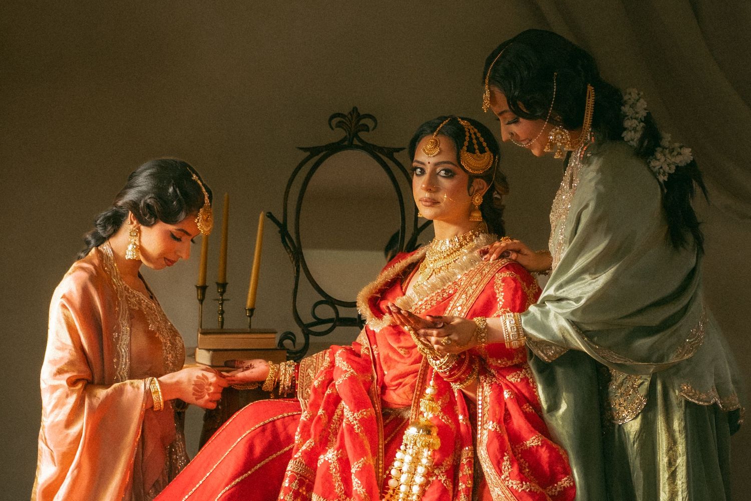 A bride in a red bridal lehenga adorned with gold jewelry is assisted by two women in traditional attire.