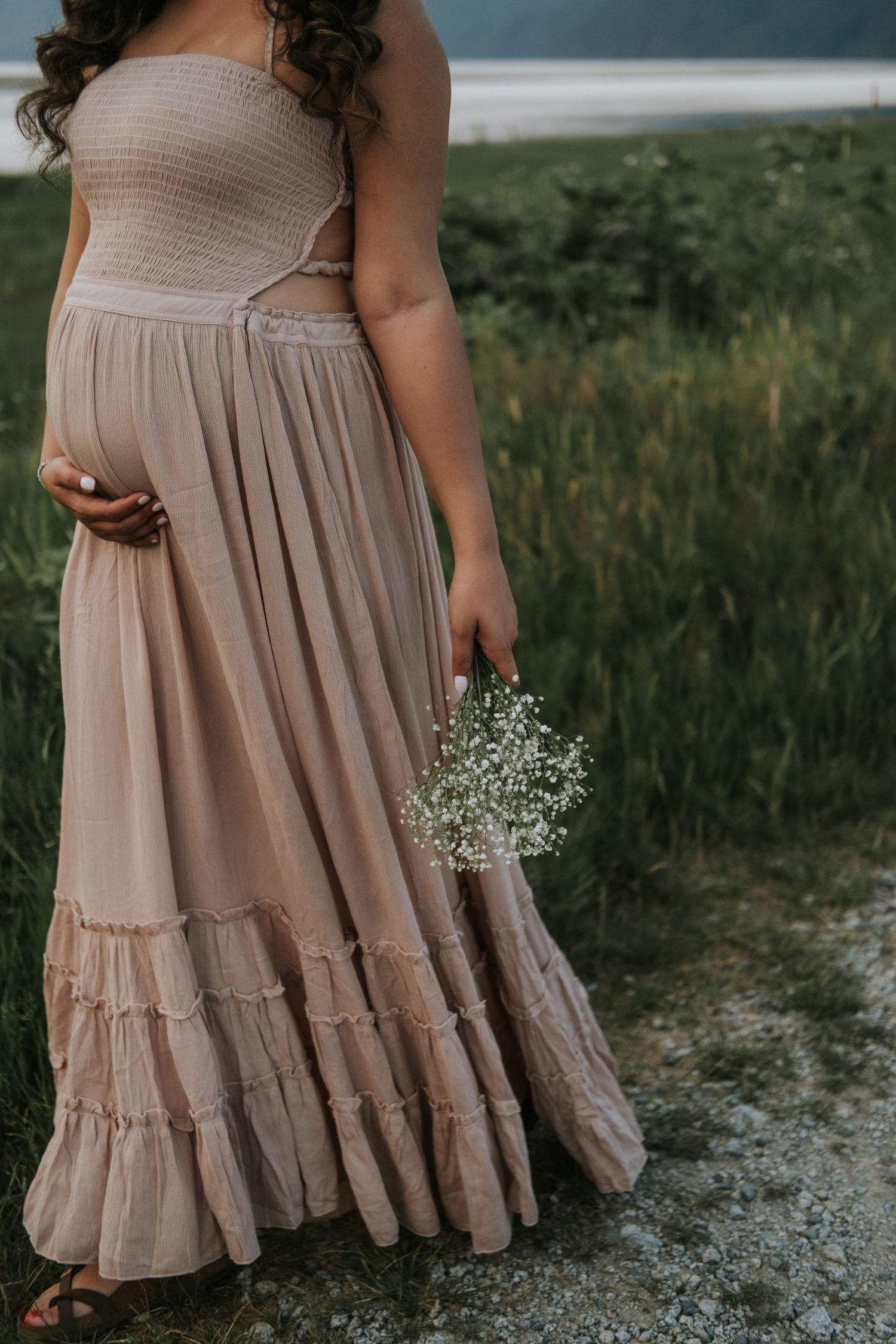 A pregnant woman in a blush strapless maxi dress holds baby's breath flowers in a scenic outdoor setting.