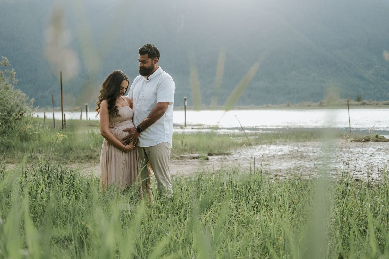 An expecting couple embraces tenderly in a lush green field beside a lake during a maternity photoshoot.
