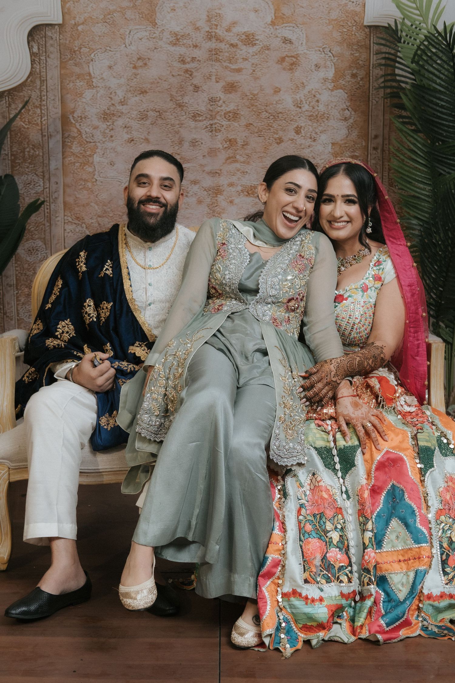 Three people in traditional South Asian attire smile joyfully while seated together at a wedding celebration.