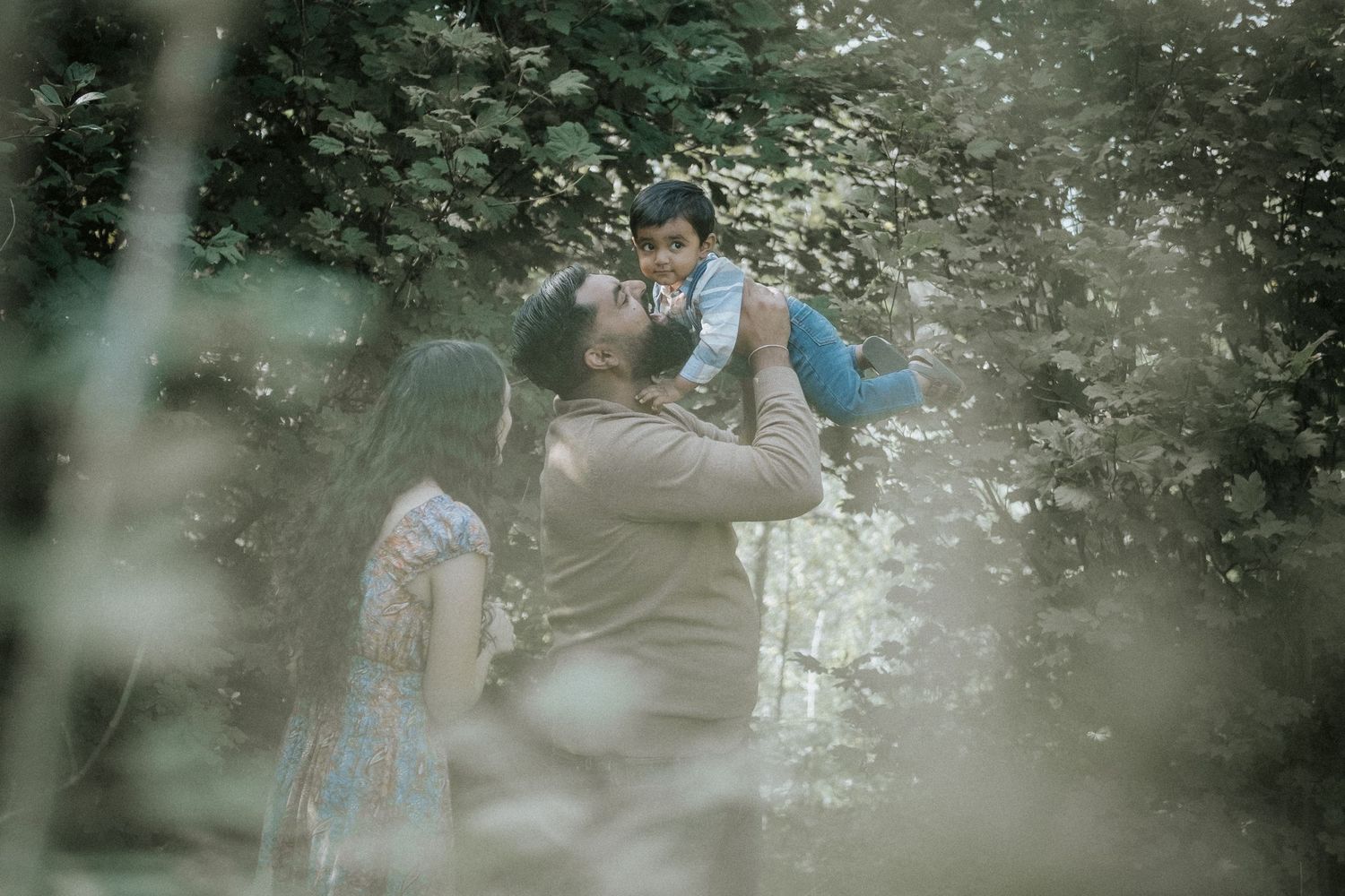 A family poses outdoors among lush green trees, with a father lifting his baby joyfully while mother stands beside them.