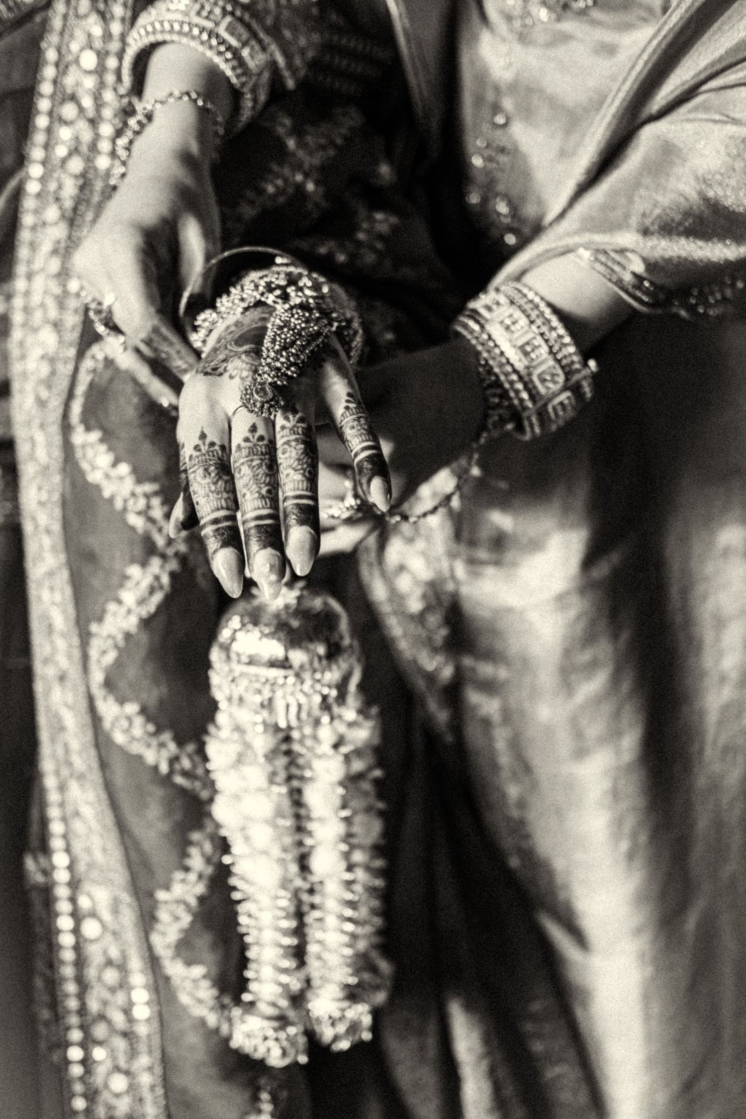 Close-up of Indian bride's hands with intricate mehndi, bangles, and a floral garland in black and white.