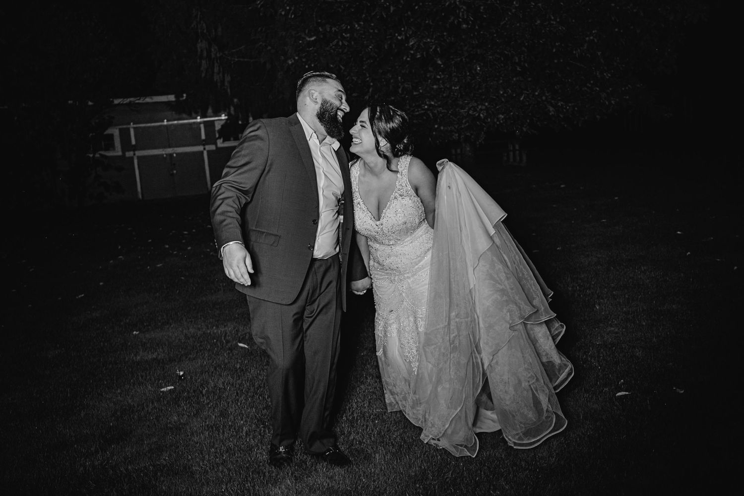 Black and white photo of newlywed couple kissing outdoors at night, groom in suit and bride in gown.