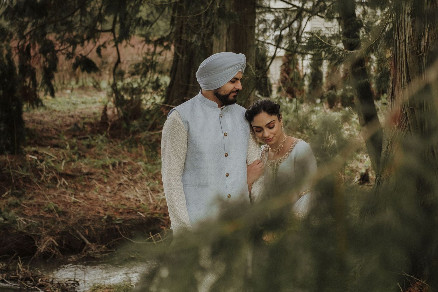 Sikh groom in white sherwani and blue turban walking with bride through lush forest wedding setting.