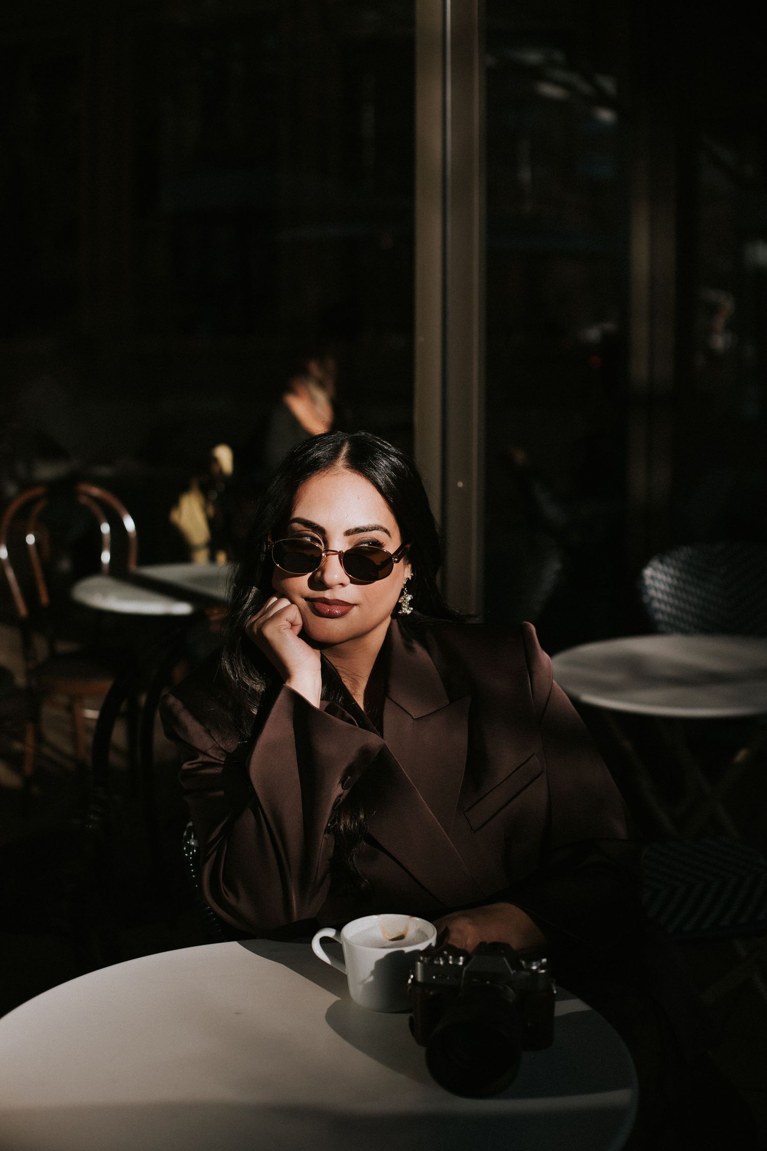 Stylish woman in brown coat and sunglasses sitting at a dark cafe table with coffee.
