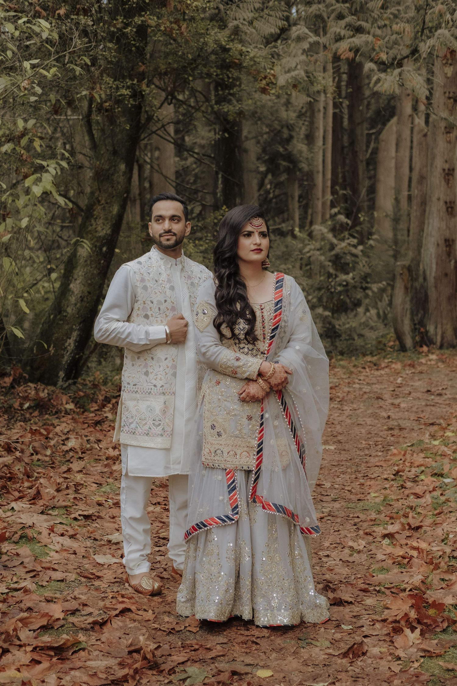 South Asian couple in matching ivory embroidered traditional outfits pose in autumn forest setting.